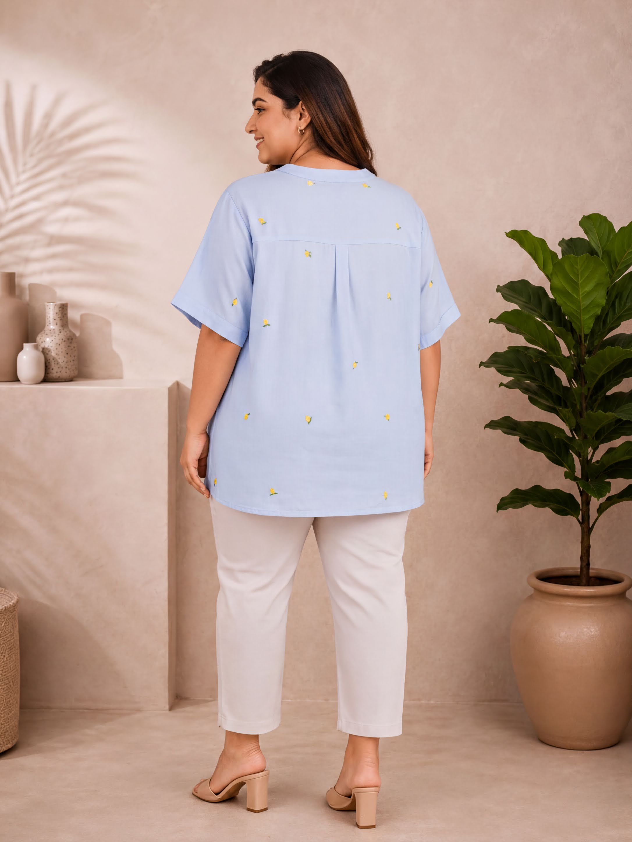 A woman wearing a light blue blouse with yellow flower designs stands in a room with a potted plant and a shelf with decorative items.