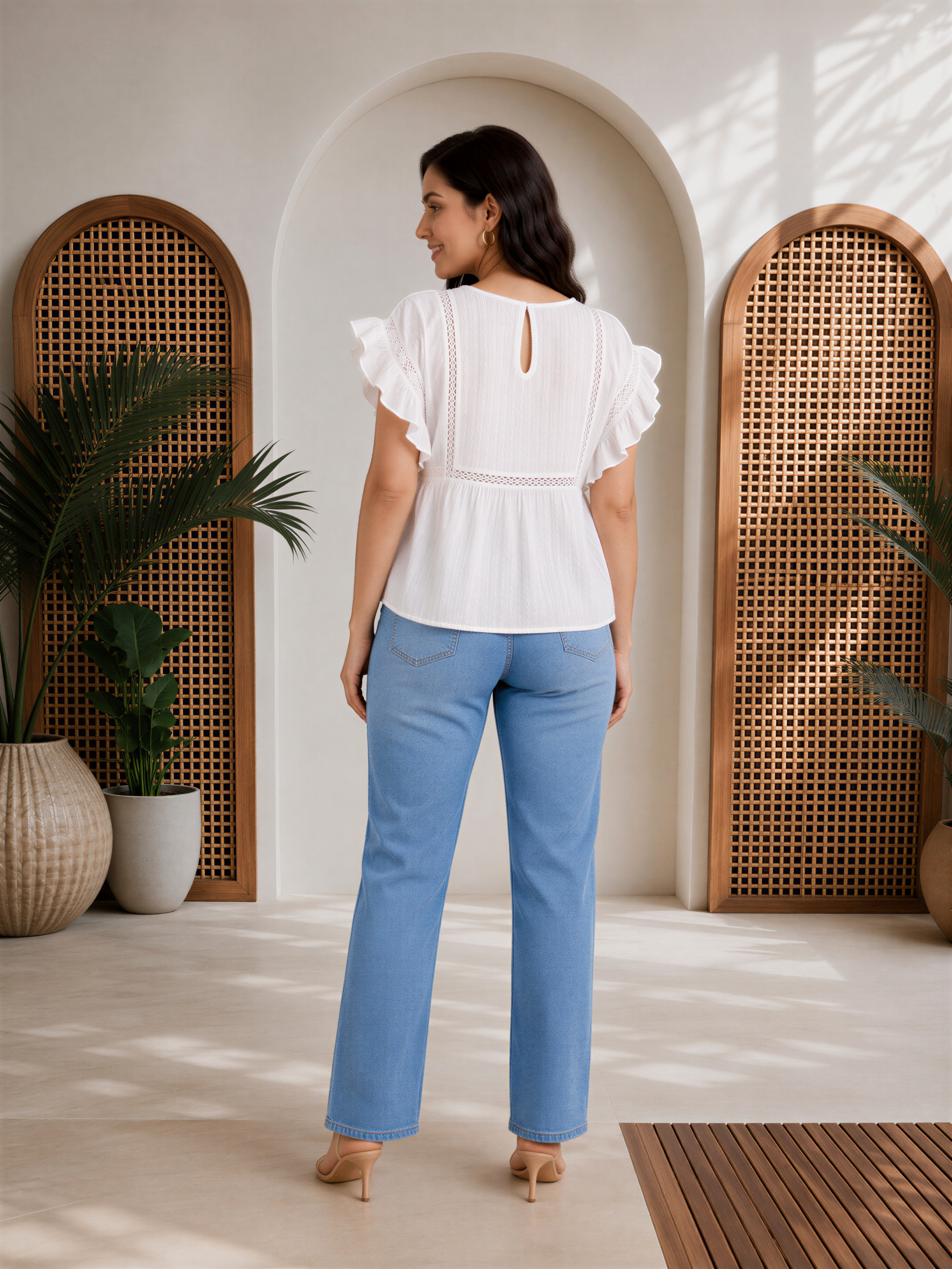 A woman wearing a white blouse with ruffled sleeves and blue jeans stands in a room with wooden arches and potted plants.