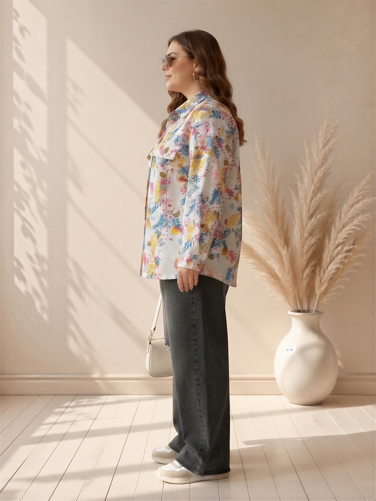 A woman wearing a floral print shirt and jeans stands in a room with a white vase of dried flowers on a table.