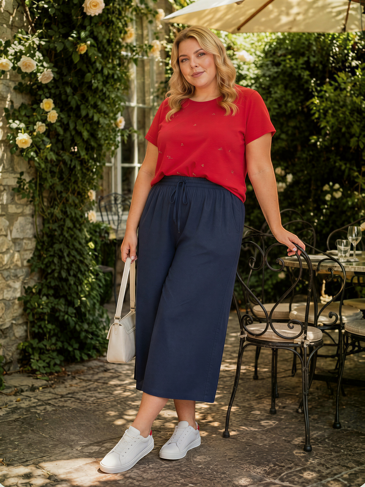 A woman in a red top and navy blue wide-leg pants stands in front of a stone wall with yellow flowers, holding a white purse and leaning against a metal chair.