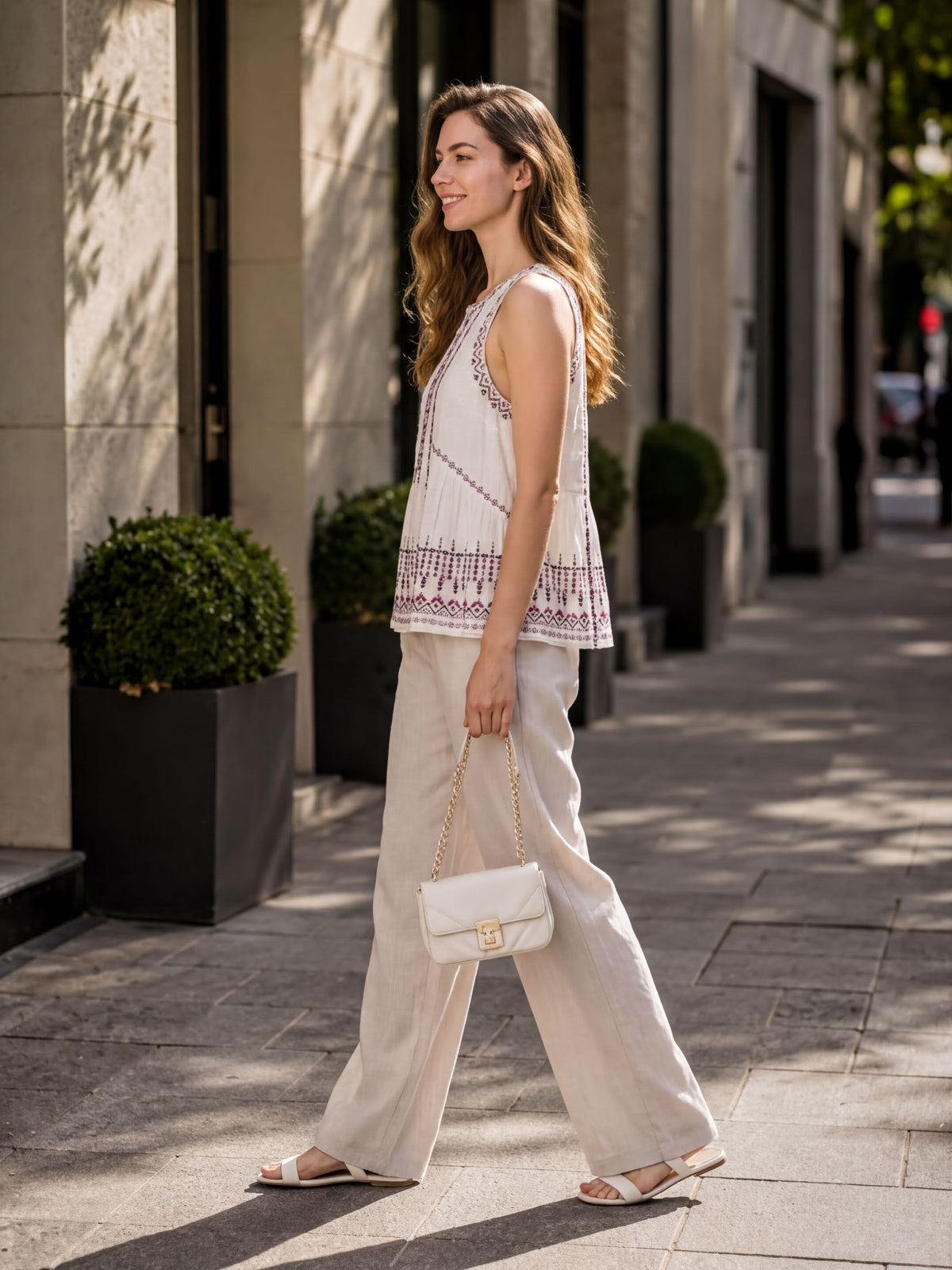 A woman wearing a white top with a patterned design and beige pants is walking down a sidewalk, carrying a white handbag.
