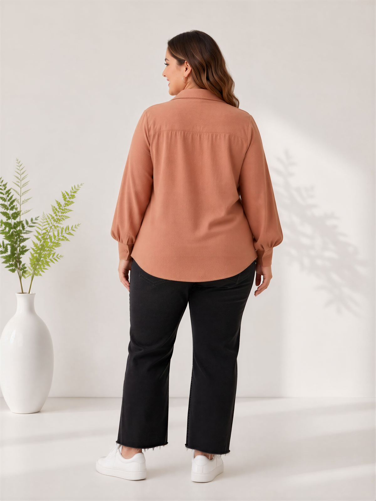 A woman wearing a peach-colored blouse and black pants stands in front of a white wall, with a potted plant to her left.