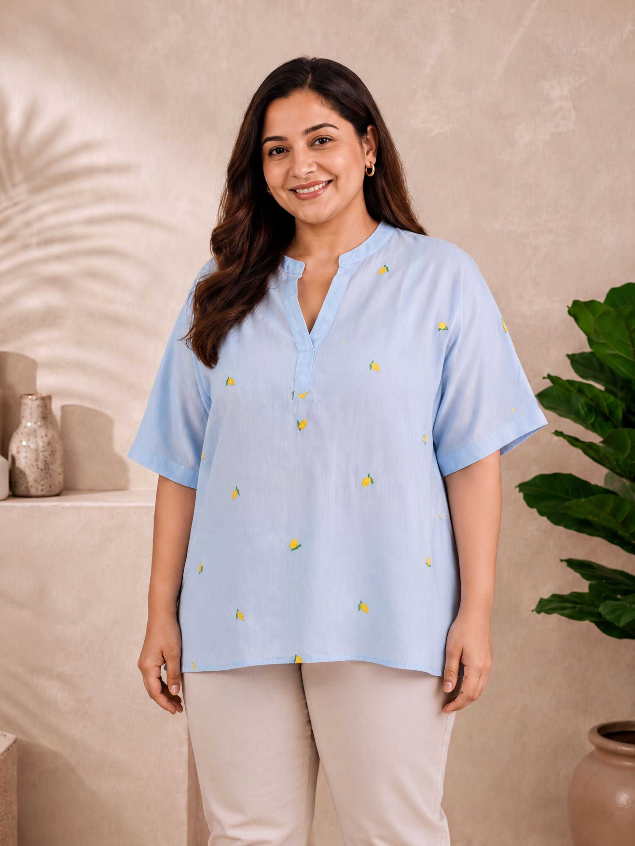 A woman wearing a light blue blouse with yellow fruit designs stands in front of a beige wall with a potted plant.