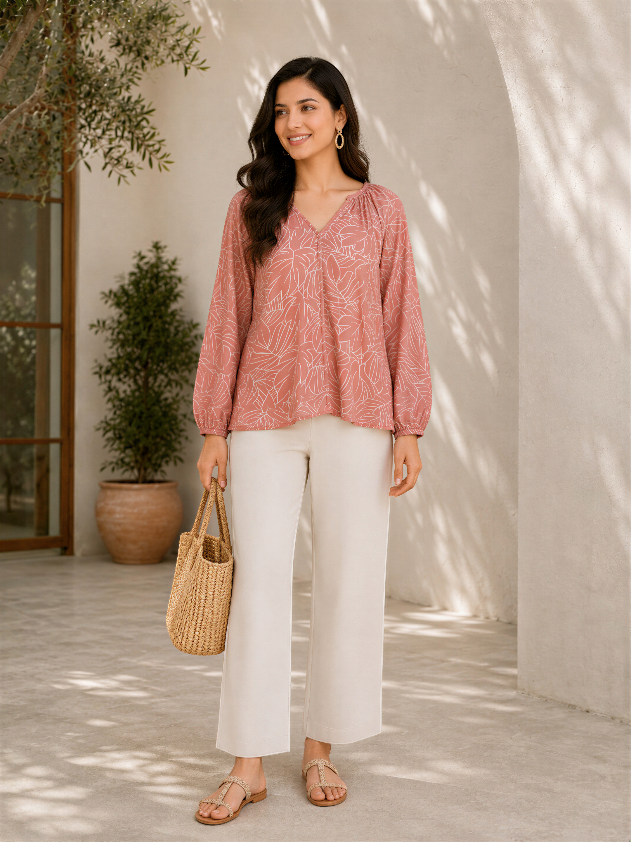 A woman wearing a pink blouse with leaf patterns, white pants, and sandals is standing in front of a white wall with a potted plant. She is holding a woven bag in her hand.