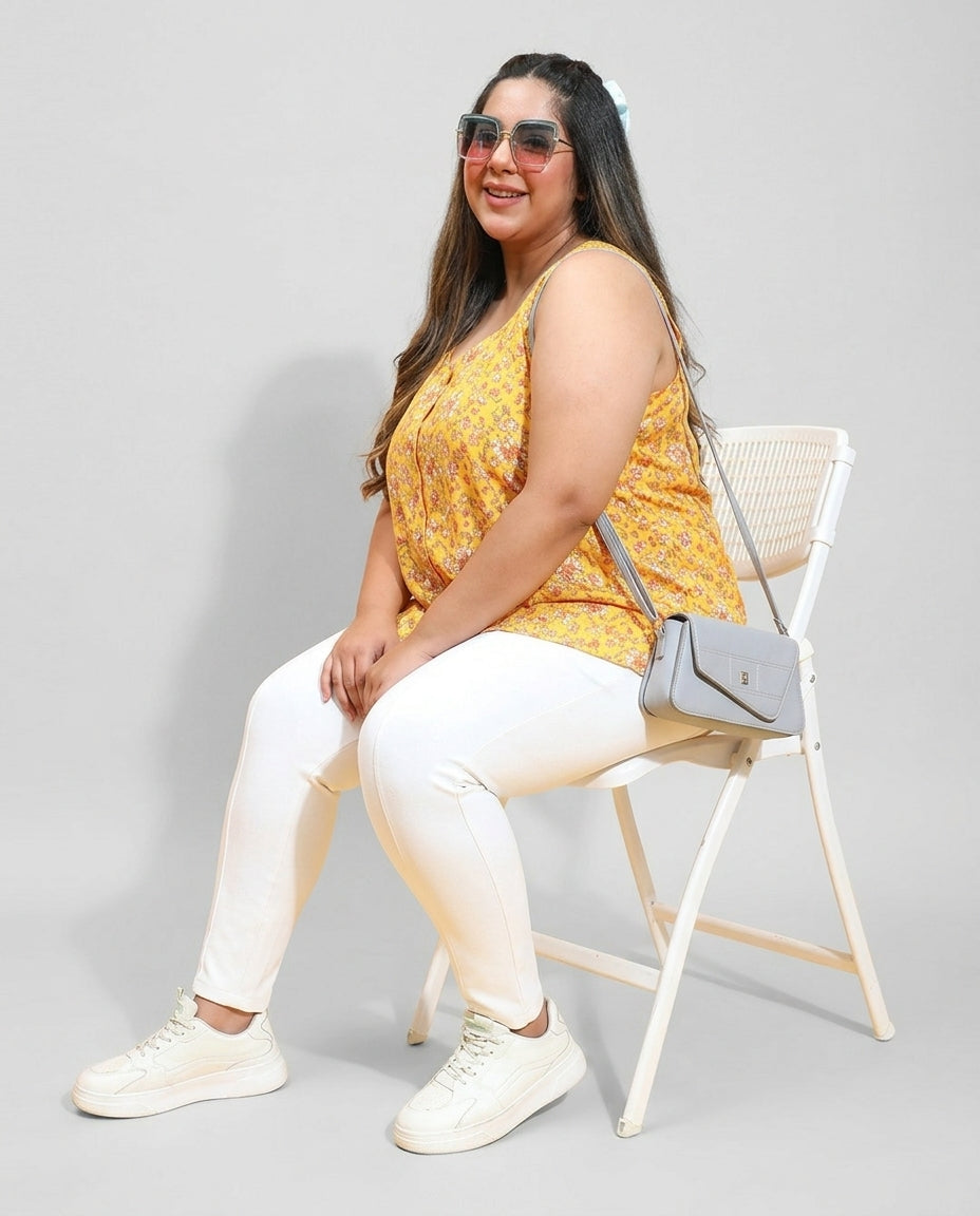 Plus size woman in yellow floral sleeveless top and white pants, sitting on chair