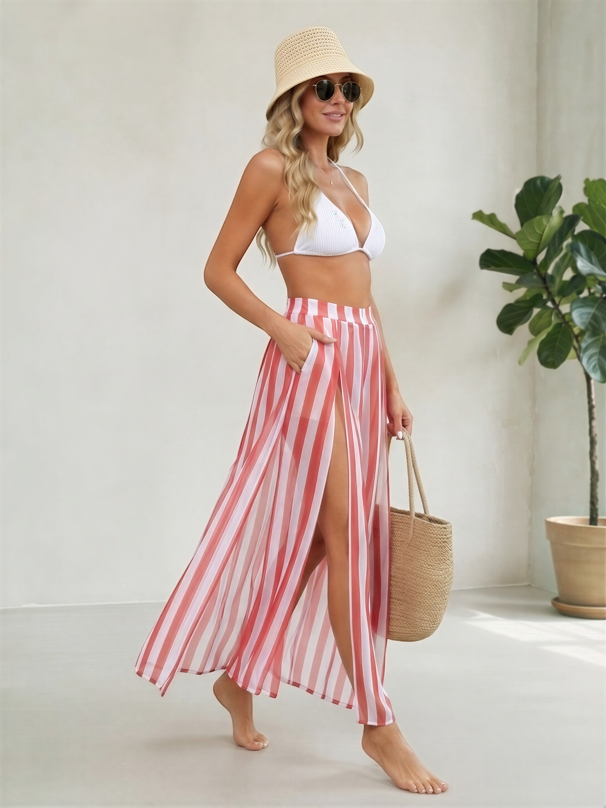 A woman wearing a white bikini top and a long, striped skirt is standing in a room with a potted plant in the background.