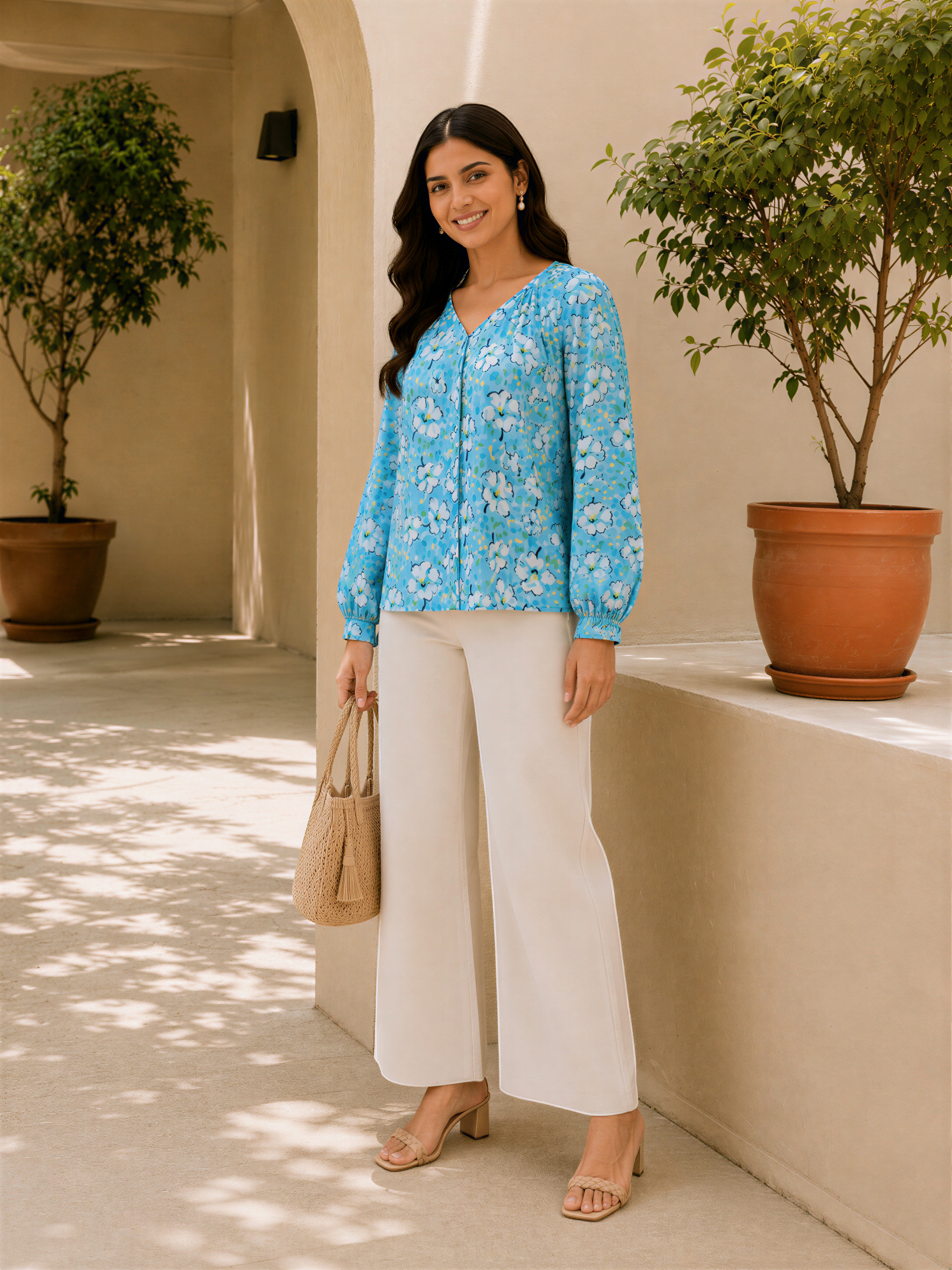 A woman wearing a blue floral blouse and white pants stands in front of a beige wall, holding a small purse.
