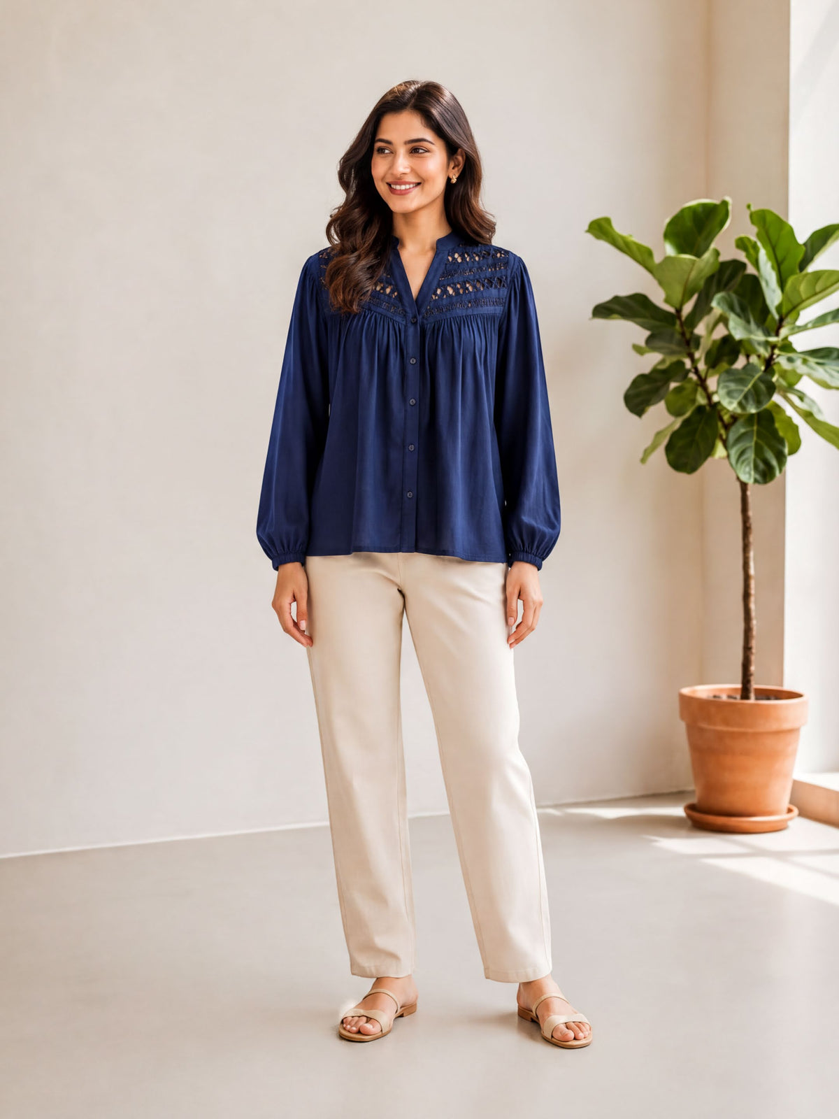 A woman wearing a navy blue blouse and beige pants stands in a room with a potted plant to her right.