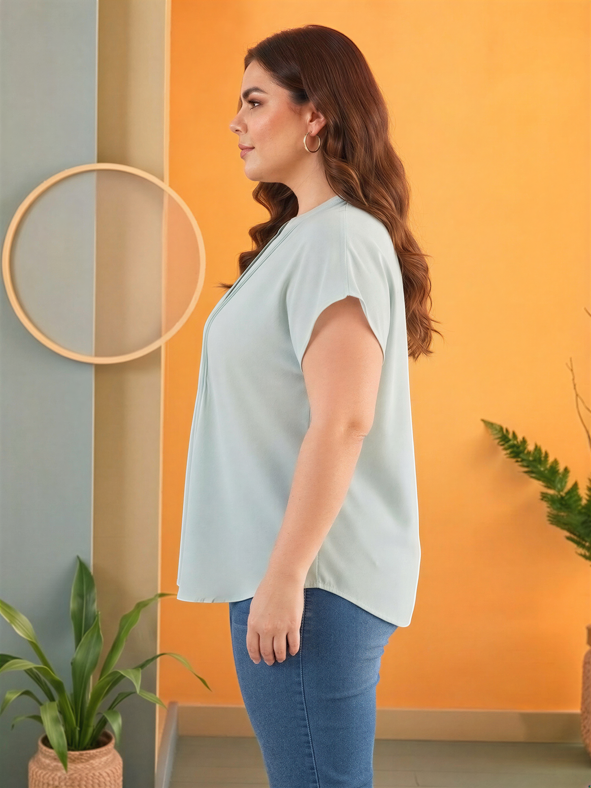 A woman with long brown hair stands in a room with a light blue top and blue jeans, wearing earrings. The room has a modern and stylish design, with a large circular mirror on the wall and potted plants in the background.