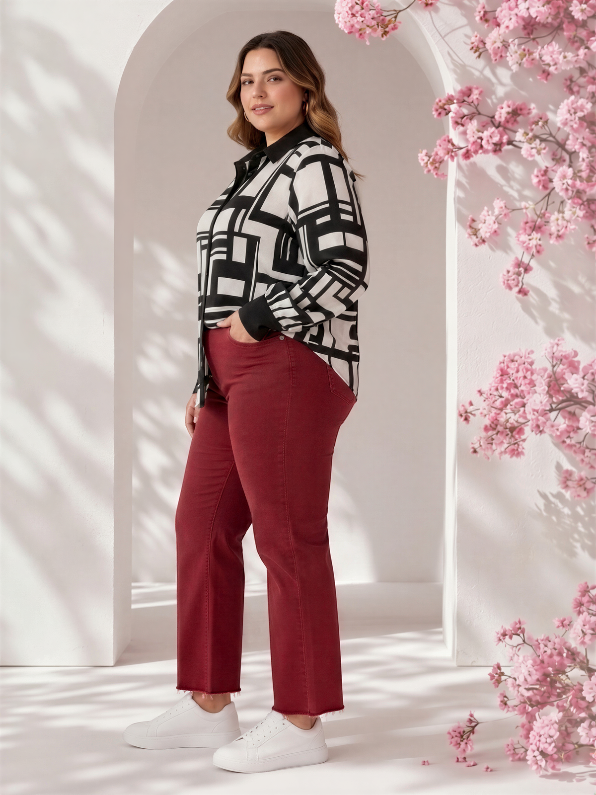 A woman wearing a black and white patterned blazer, red pants, and white sneakers stands in front of a white archway with pink flowers.
