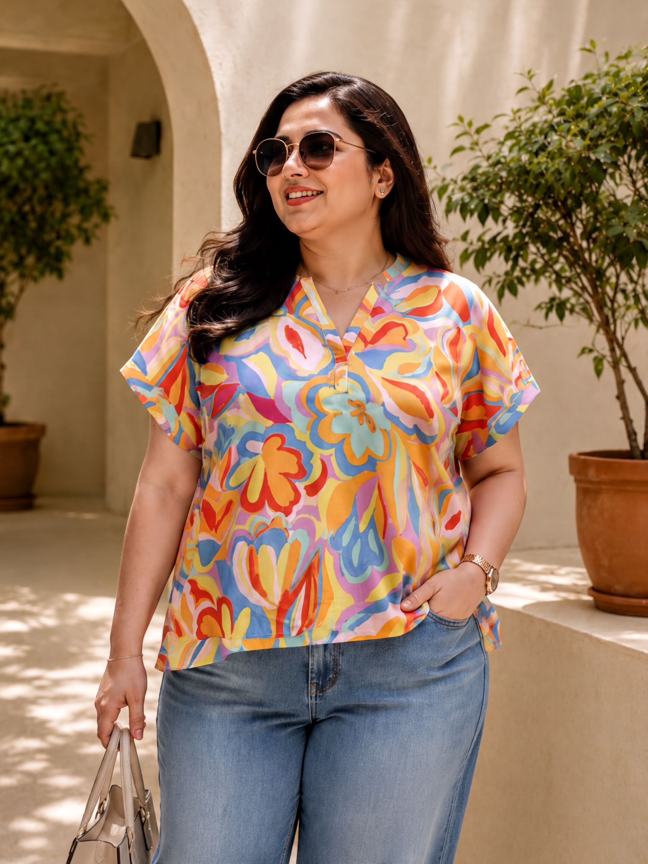 A woman wearing a colorful floral top and sunglasses stands in front of a building with potted plants.