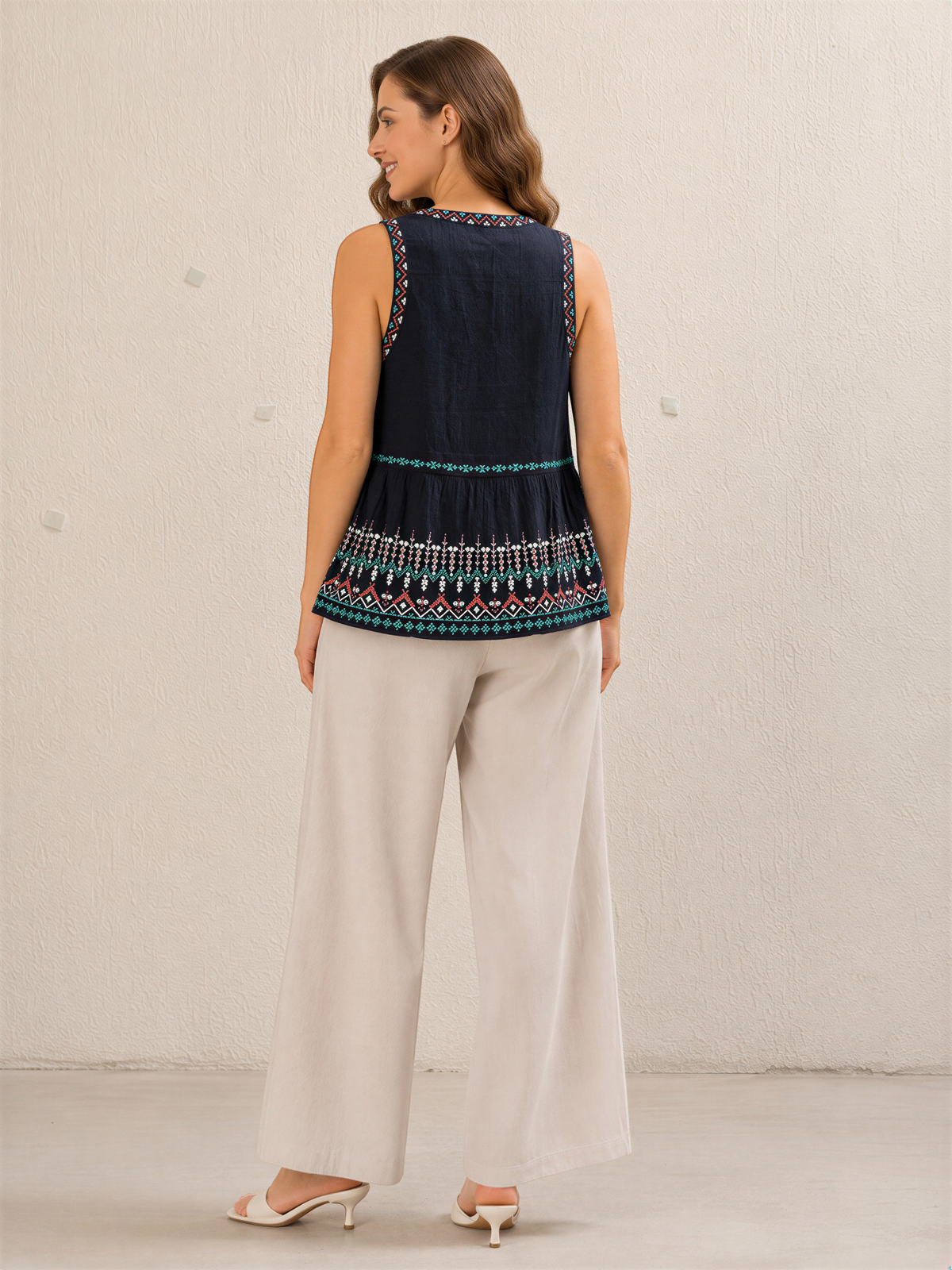 A woman wearing a navy blue sleeveless top with a patterned design and beige wide-leg pants stands against a plain white wall.