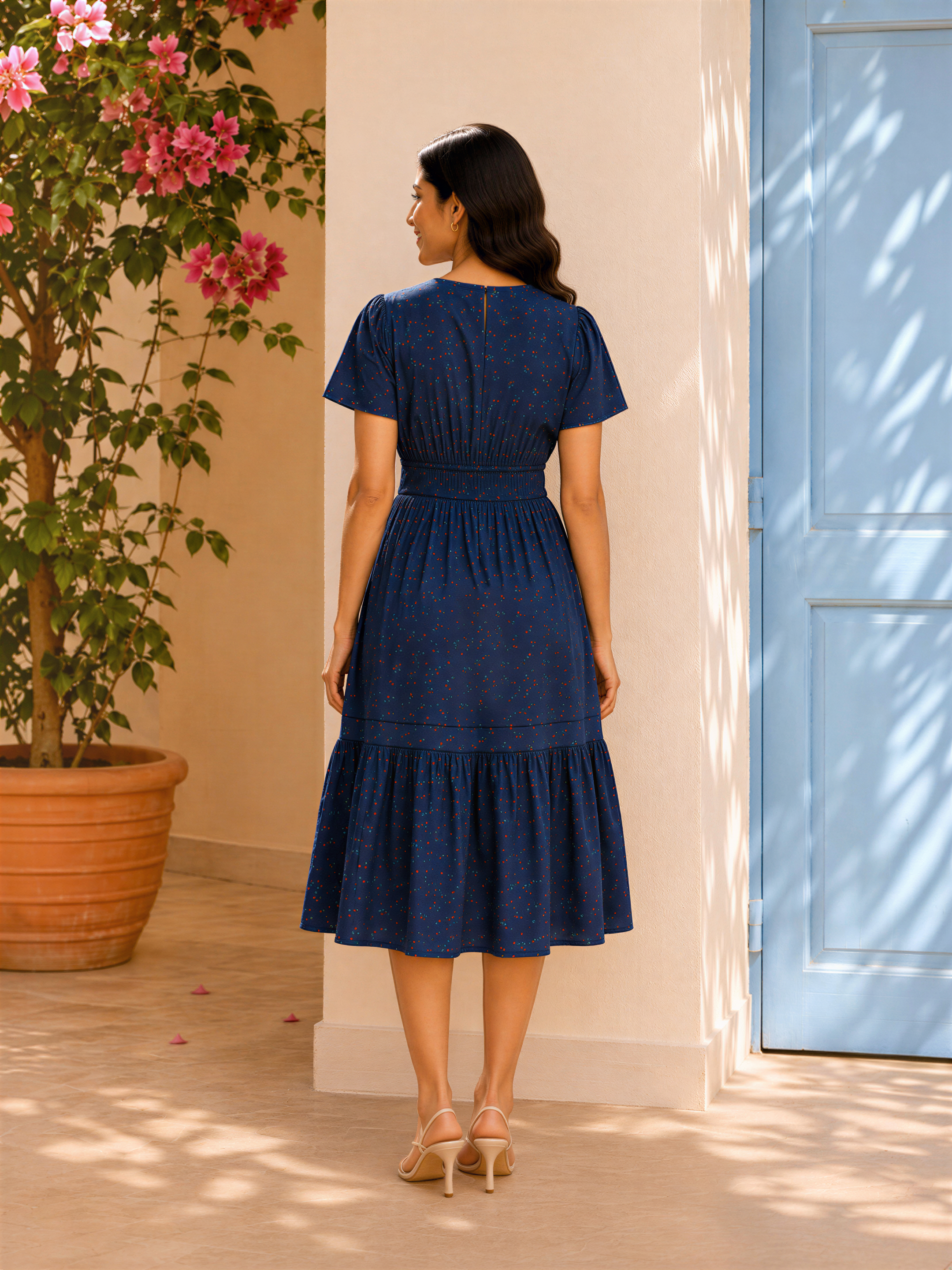 A woman in a blue dress stands in front of a blue door, with pink flowers in a pot to her left.