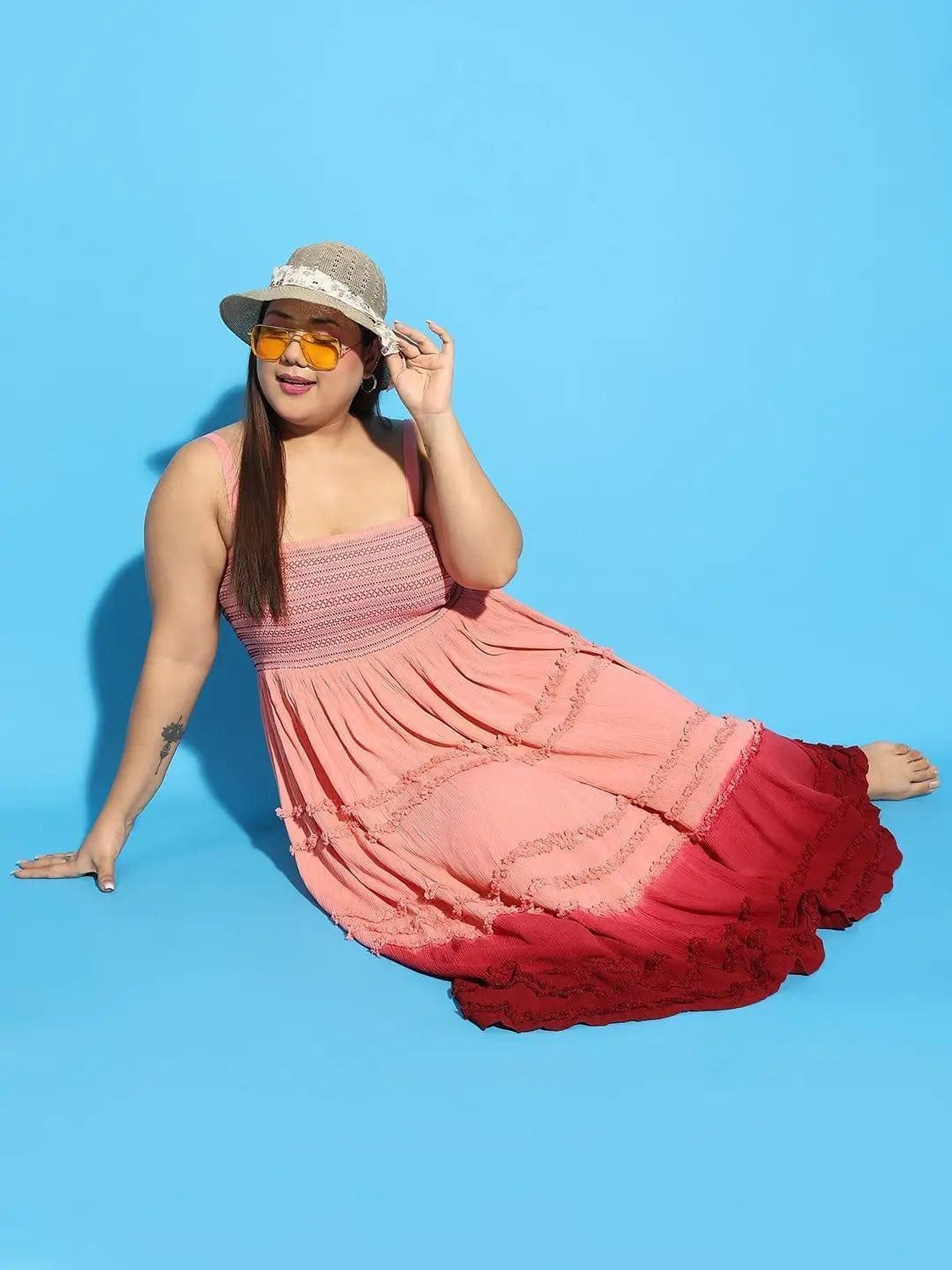 A woman wearing a pink and red tiered dress and a wide-brimmed hat is smiling and gesturing with her hand.