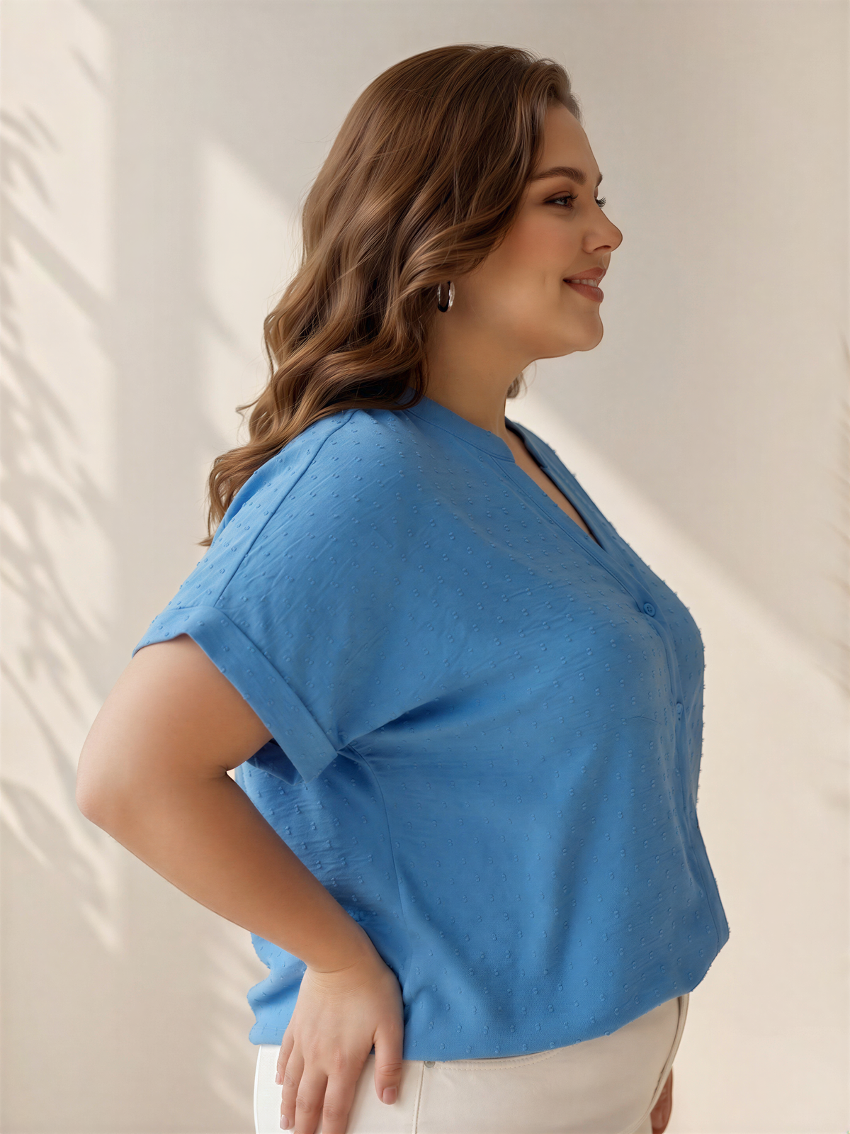A woman with long, wavy brown hair wearing a blue blouse stands against a plain white background, smiling and looking to the side.