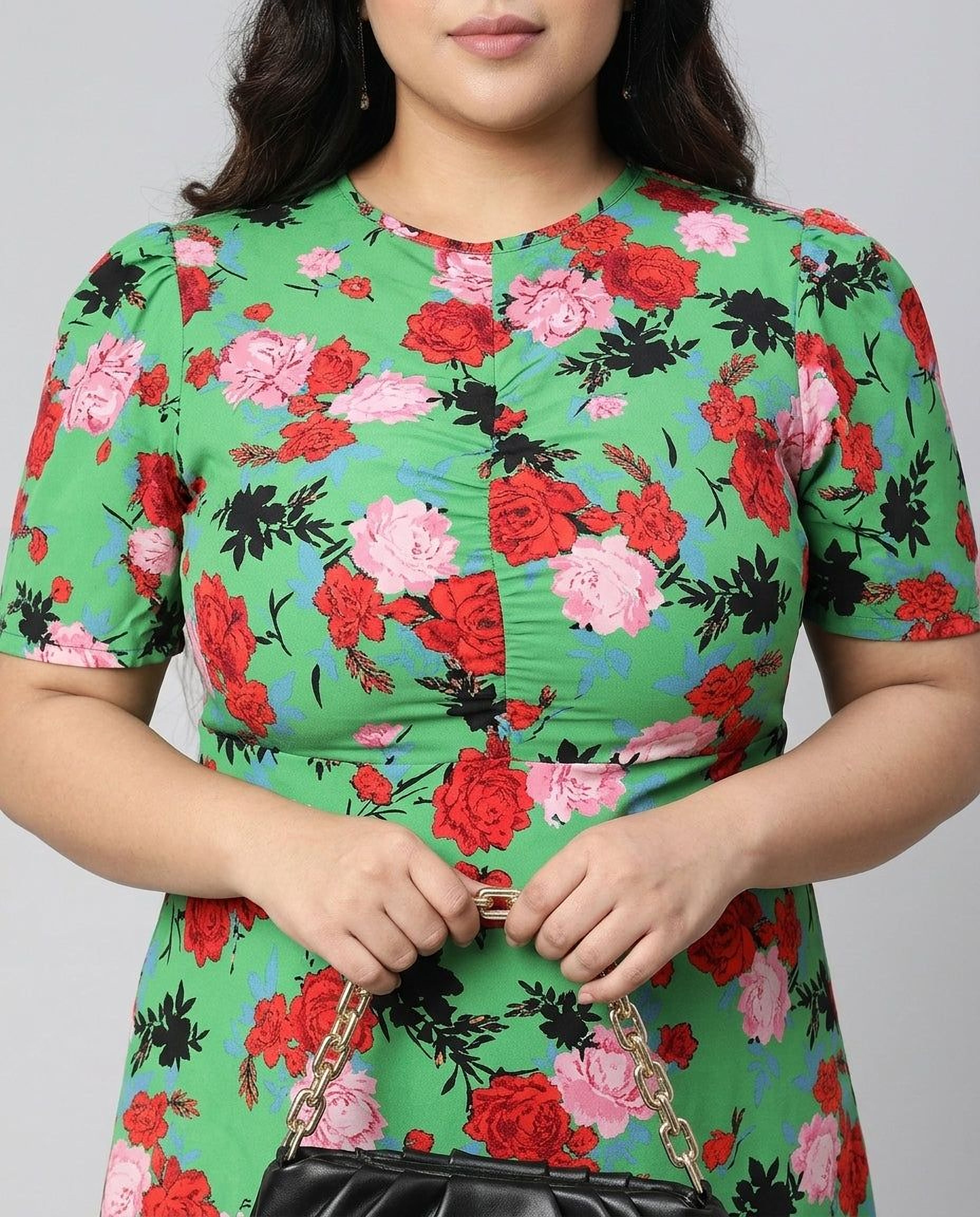 A woman wearing a green floral print dress and sunglasses, holding a black purse, and smiling at the camera.