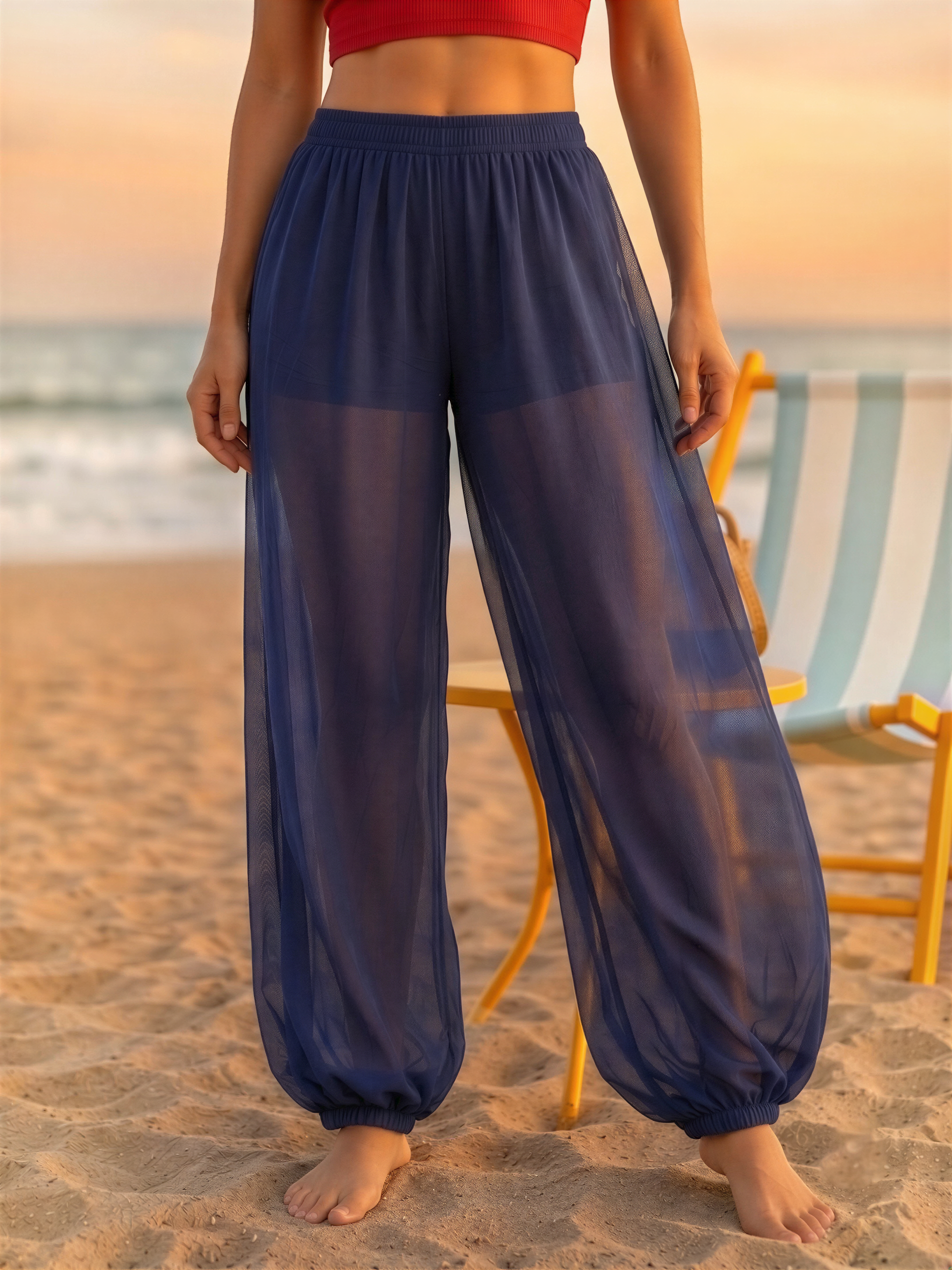 A woman wearing a red top and blue sheer pants stands on a sandy beach, with a yellow beach chair visible in the background.