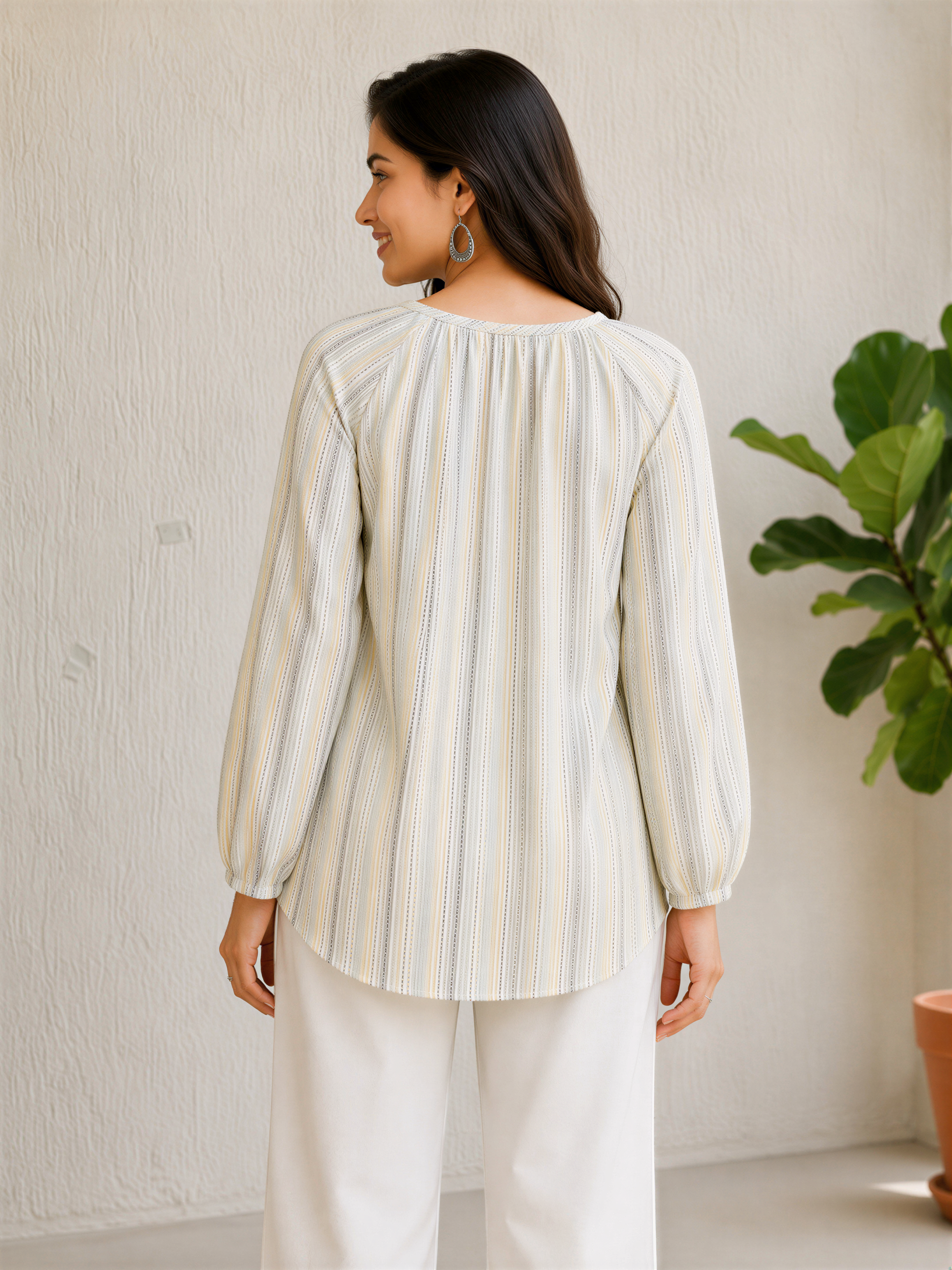 A woman wearing a striped blouse and white pants stands in front of a plain white wall, with a potted plant to her right.
