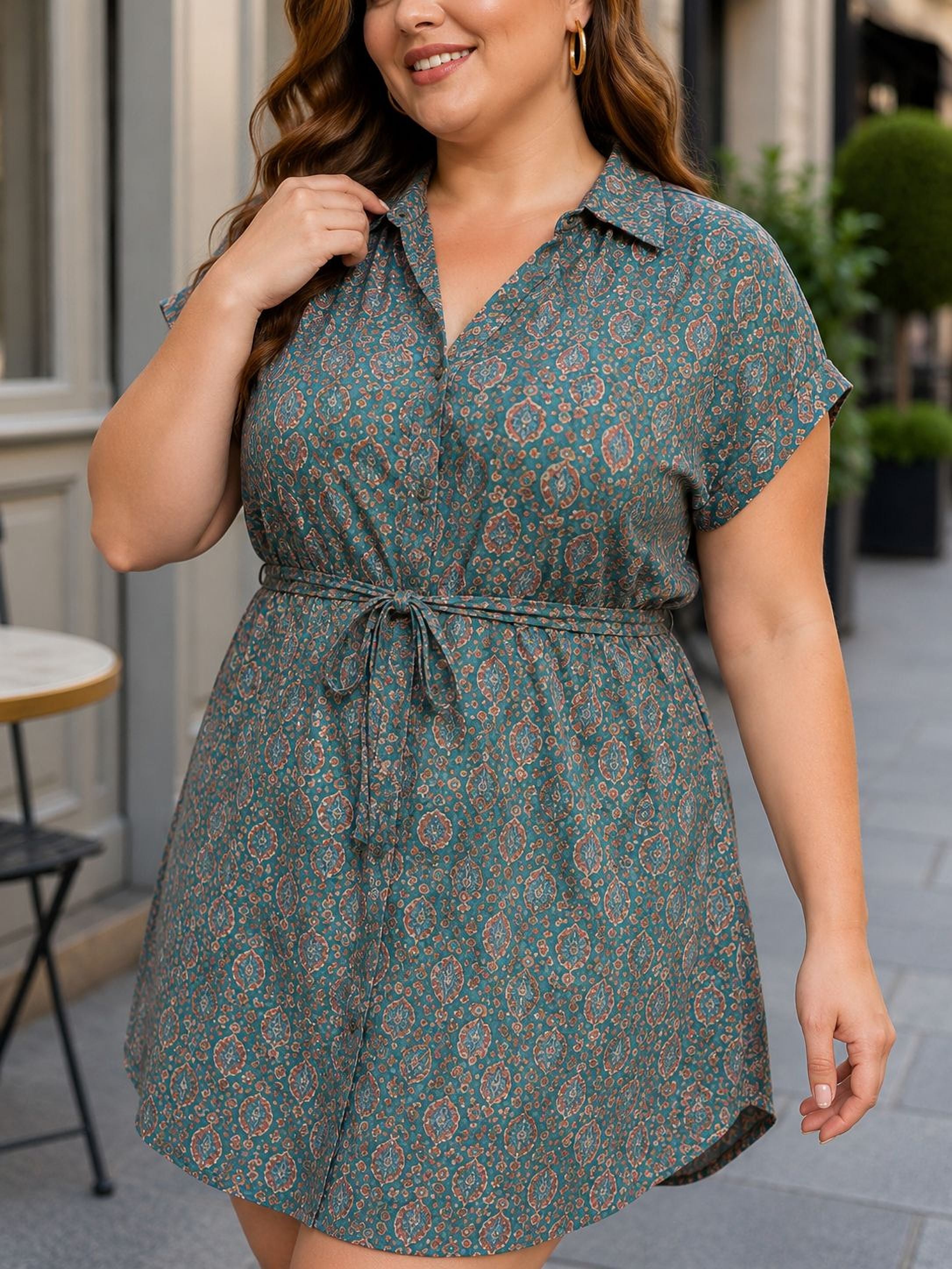 A woman wearing a green and orange patterned dress with a tie waist, smiling and walking on a sidewalk.