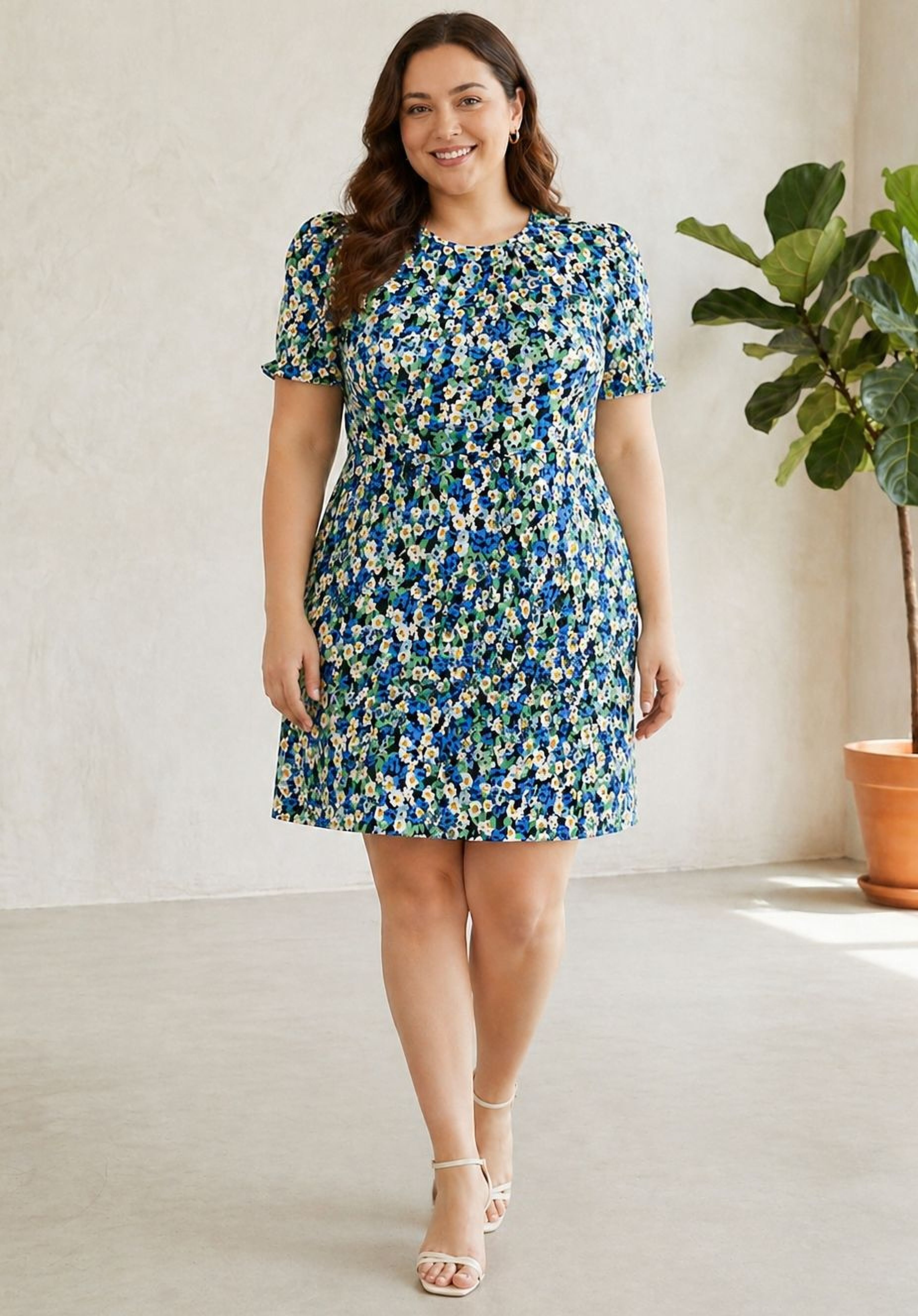 Woman wearing a blue floral dress standing in a bright room with a plant.