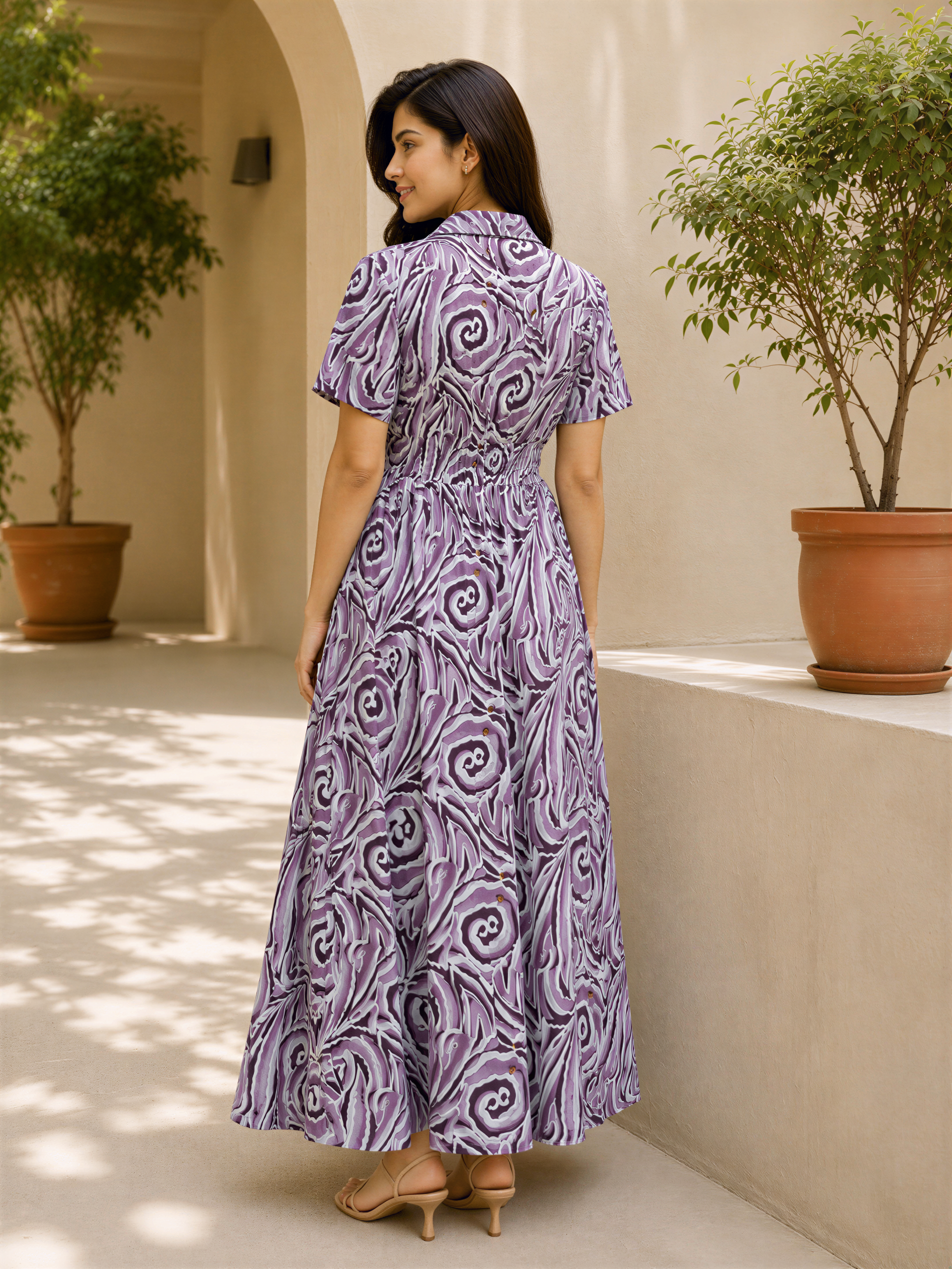 A woman wearing a long purple dress with a swirling pattern stands in front of a beige wall with potted plants.