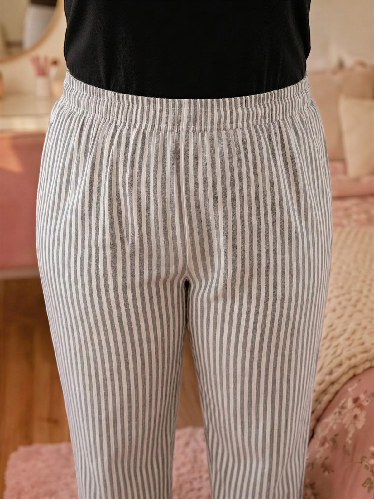 A person wearing striped pants stands in a room with a pink bedspread and a mirror in the background.