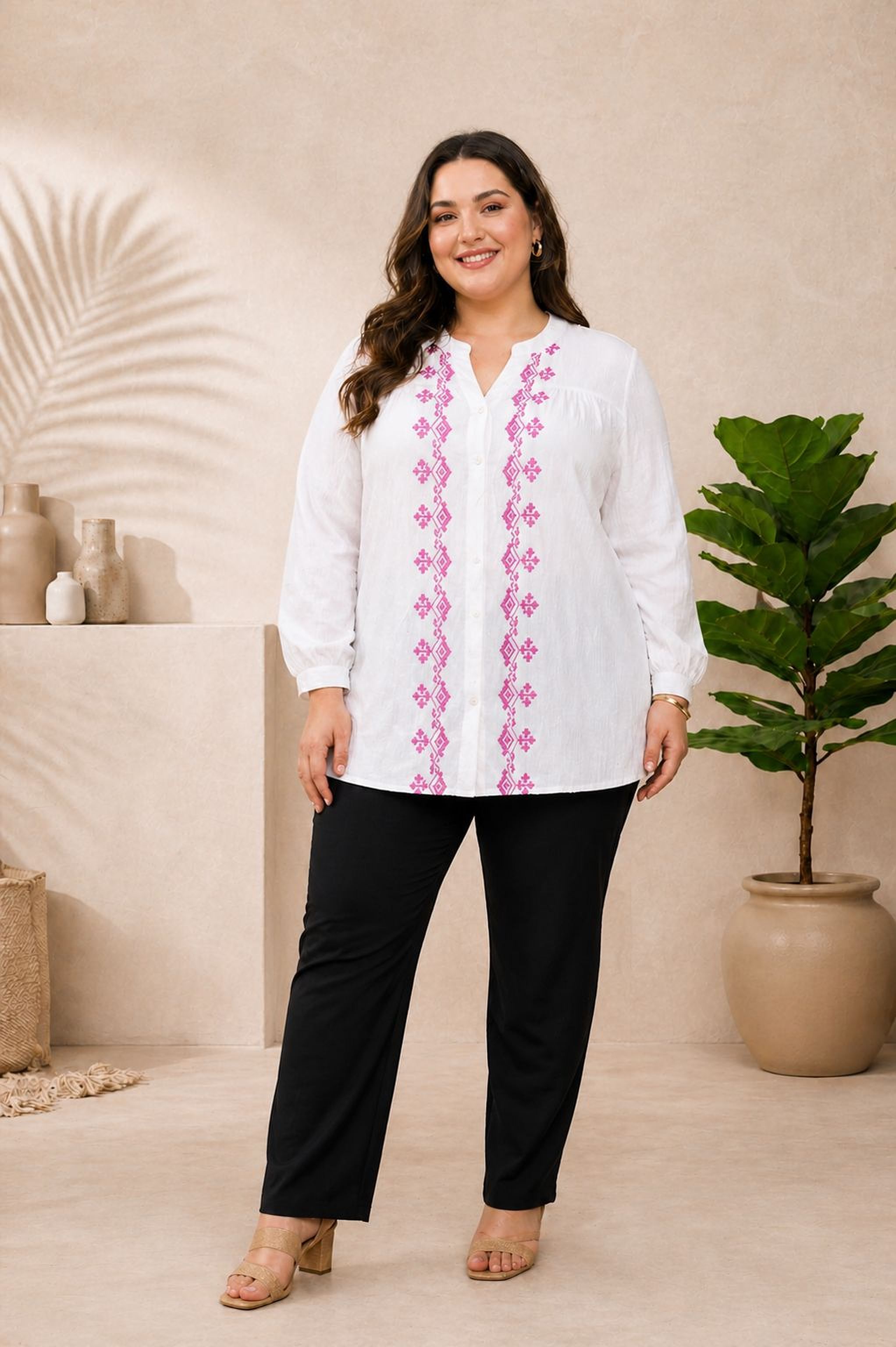 A woman wearing a white blouse with pink embroidered designs and black pants stands in front of a beige wall with a potted plant.