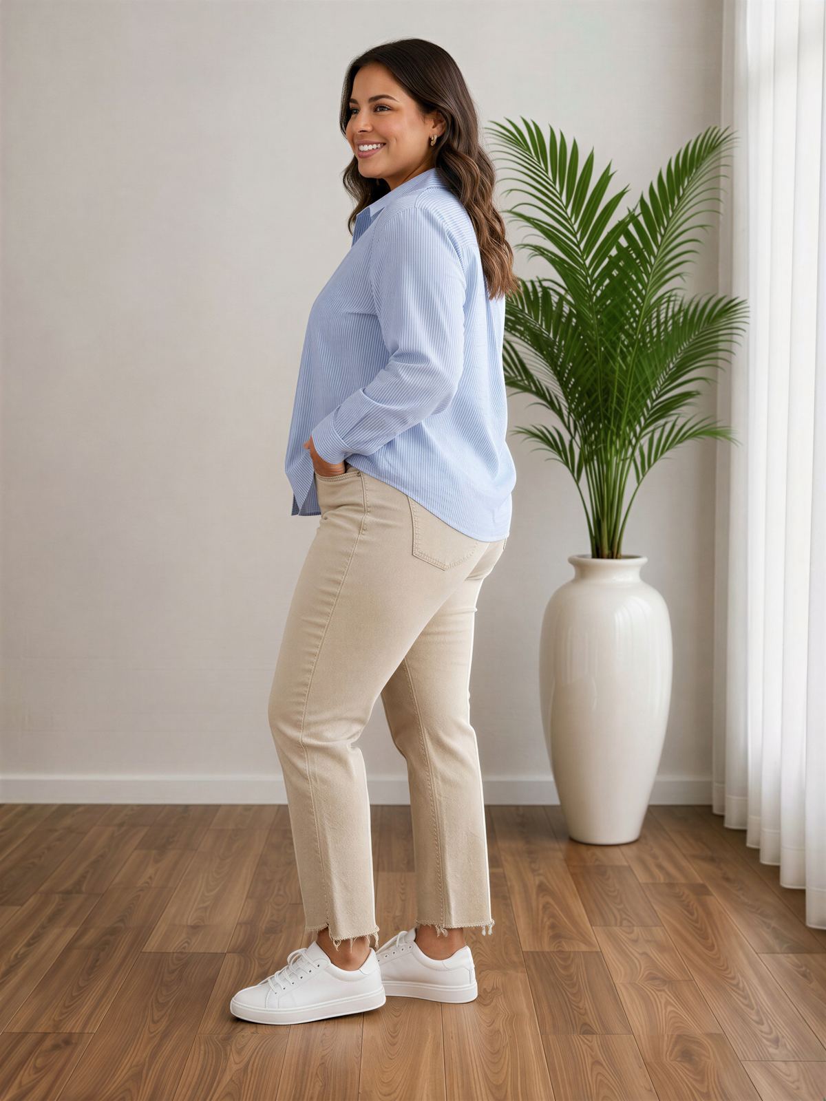 A woman in a blue shirt and beige pants stands in a room with a large potted plant and a wooden floor.