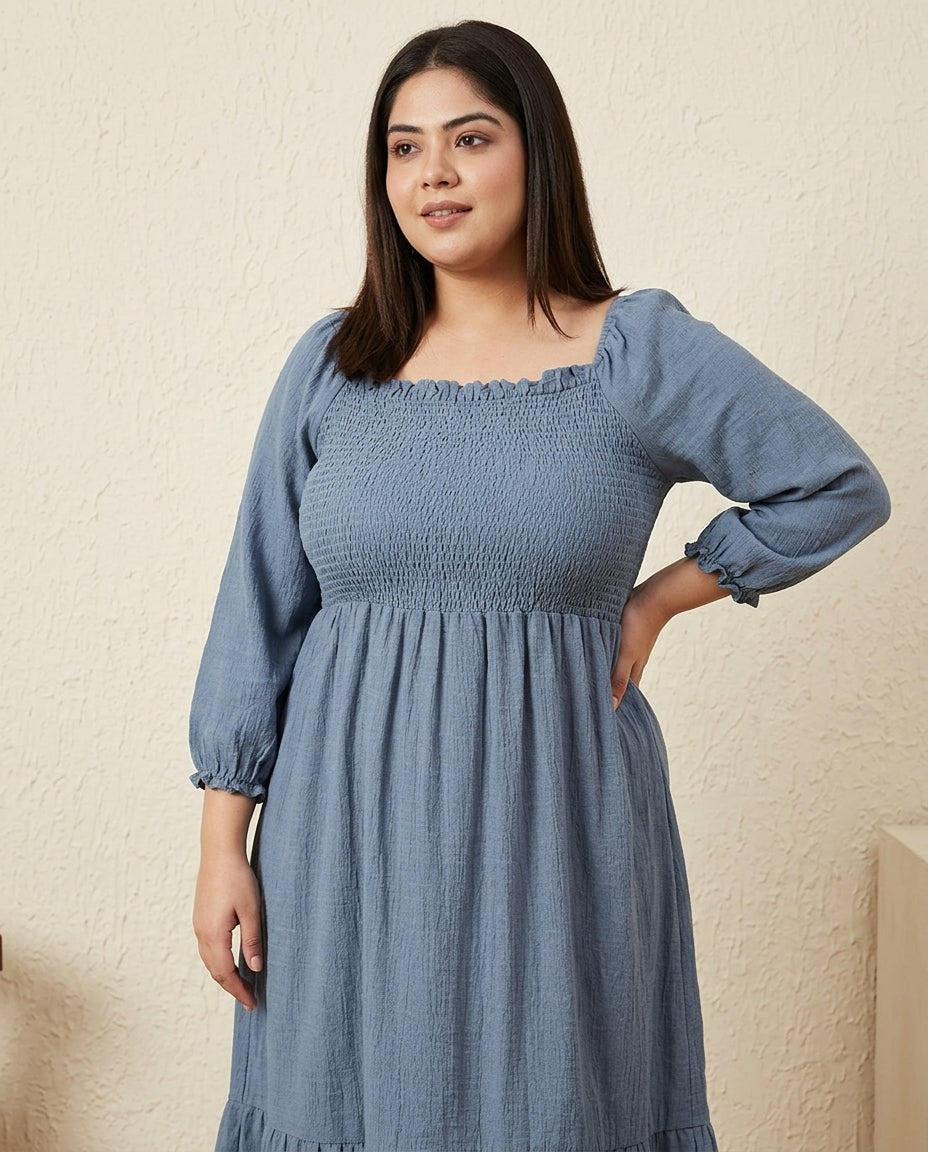 Plus size woman in a blue smocked midi dress with long sleeves, standing indoors