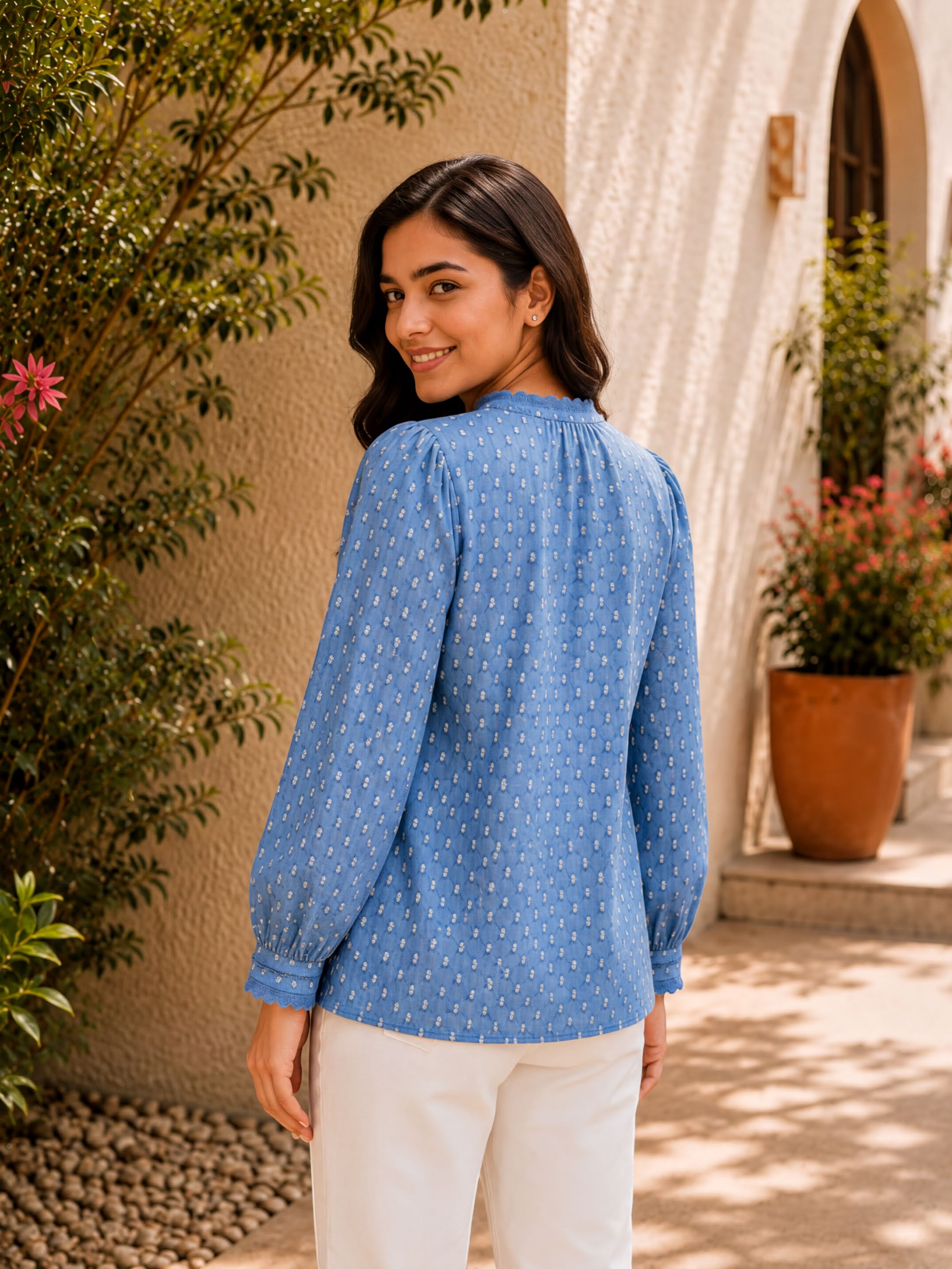 A woman wearing a blue blouse with white polka dots stands in front of a building with a pink flower, smiling at the camera.
