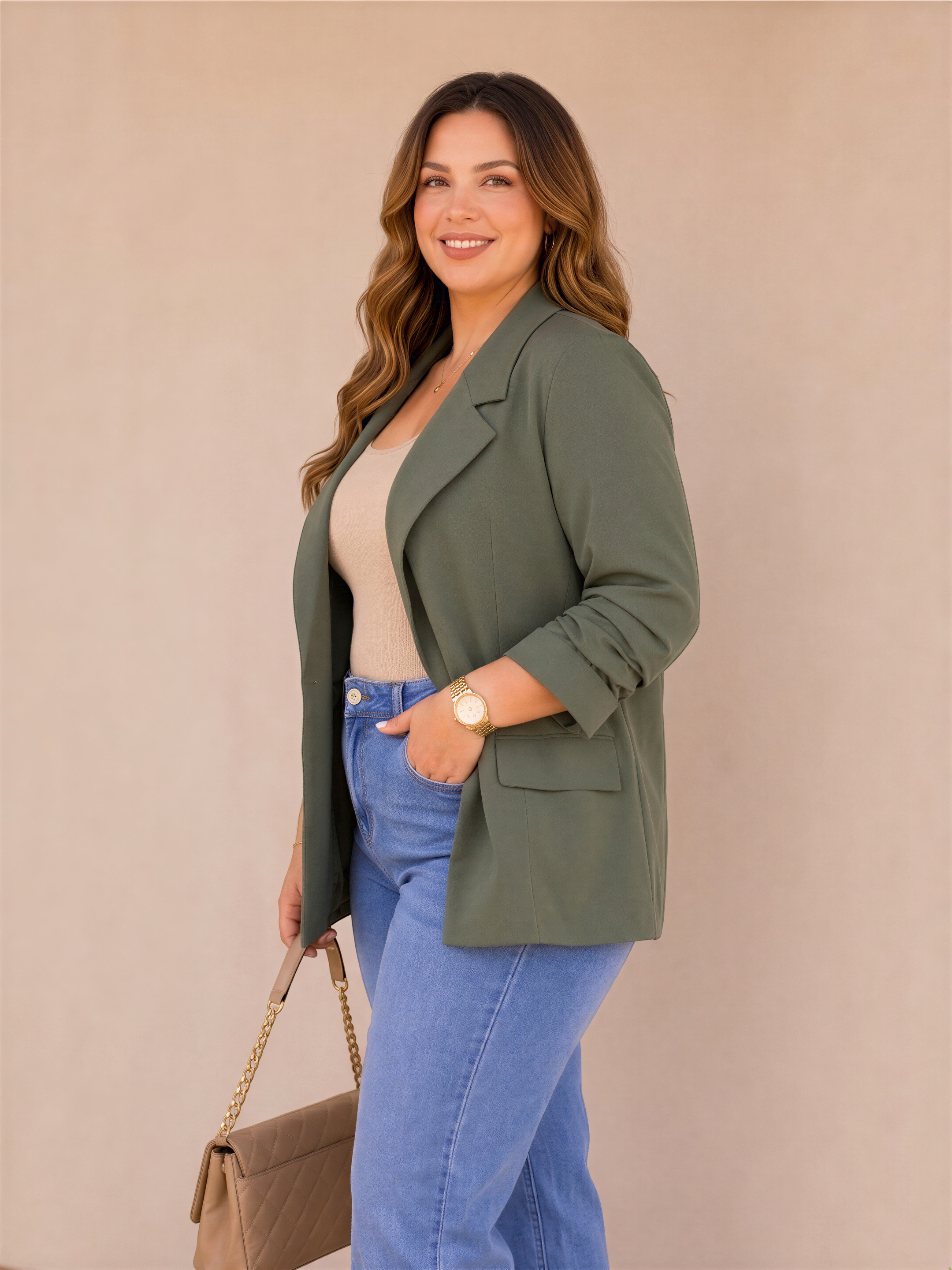 A woman with long brown hair is standing against a beige wall, wearing a green blazer, blue jeans, and a gold watch. She is smiling and holding a brown purse.