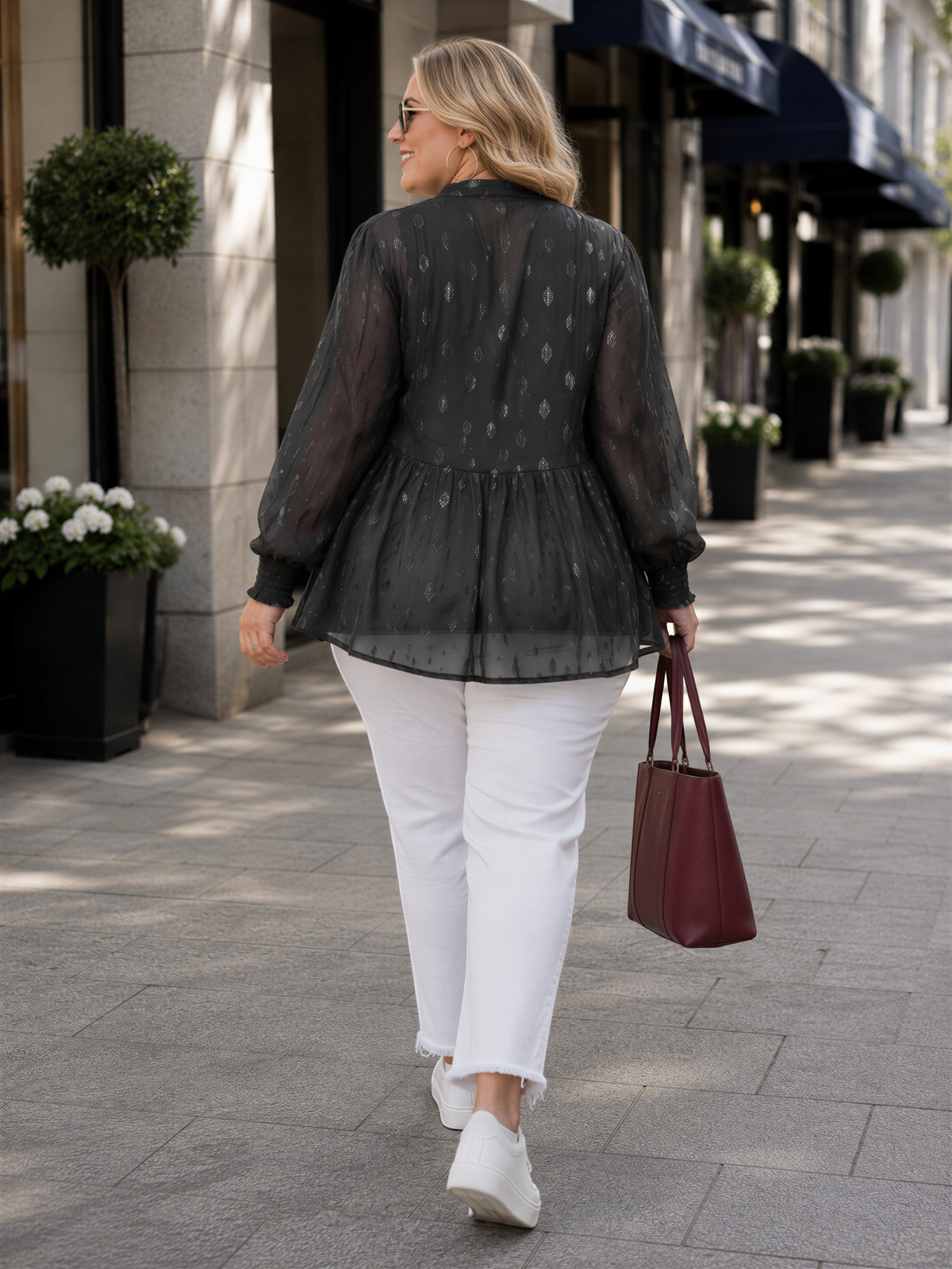 A woman wearing a black blouse with a ruffled collar and white pants is walking down a sidewalk, carrying a red handbag.