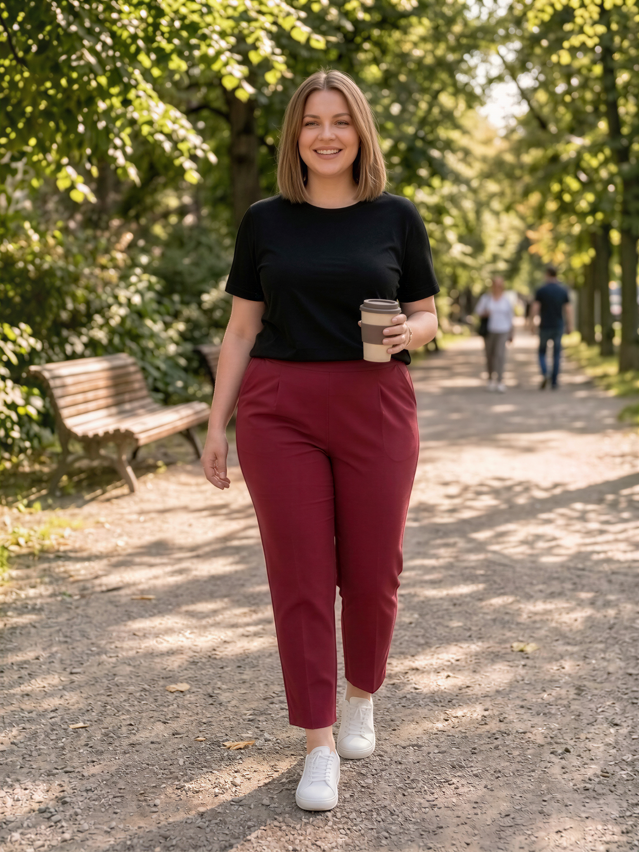 A woman in a black top and red pants is walking on a path in a park, holding a coffee cup.