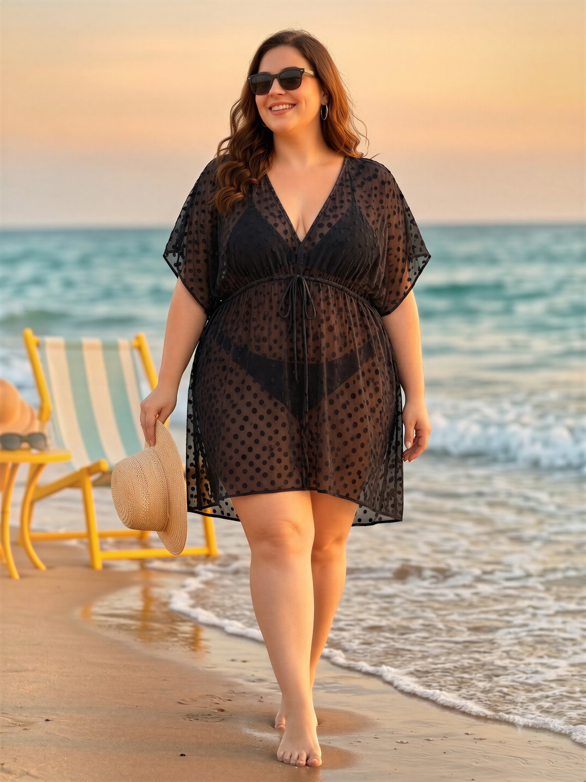A woman in a black polka dot dress is walking on a beach, holding a straw hat and smiling.