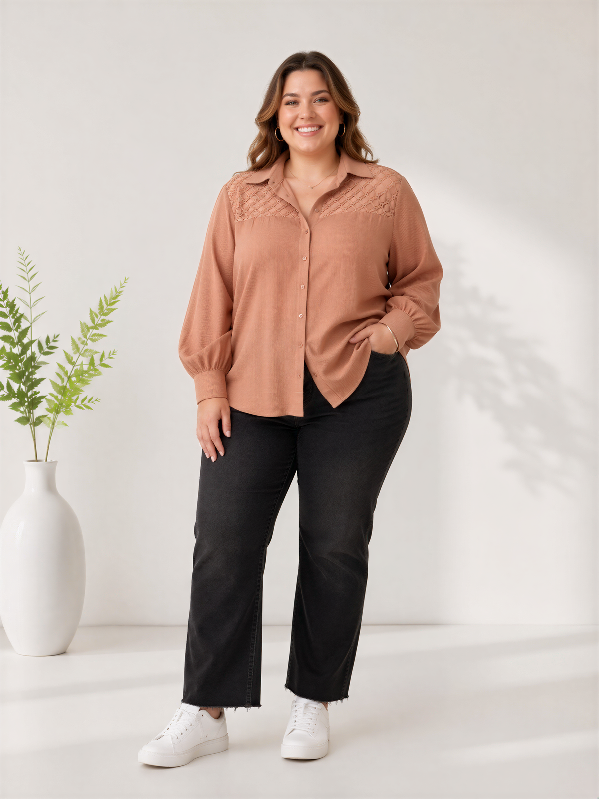 A woman wearing a peach-colored blouse and black pants stands in a minimalist setting, smiling at the camera.