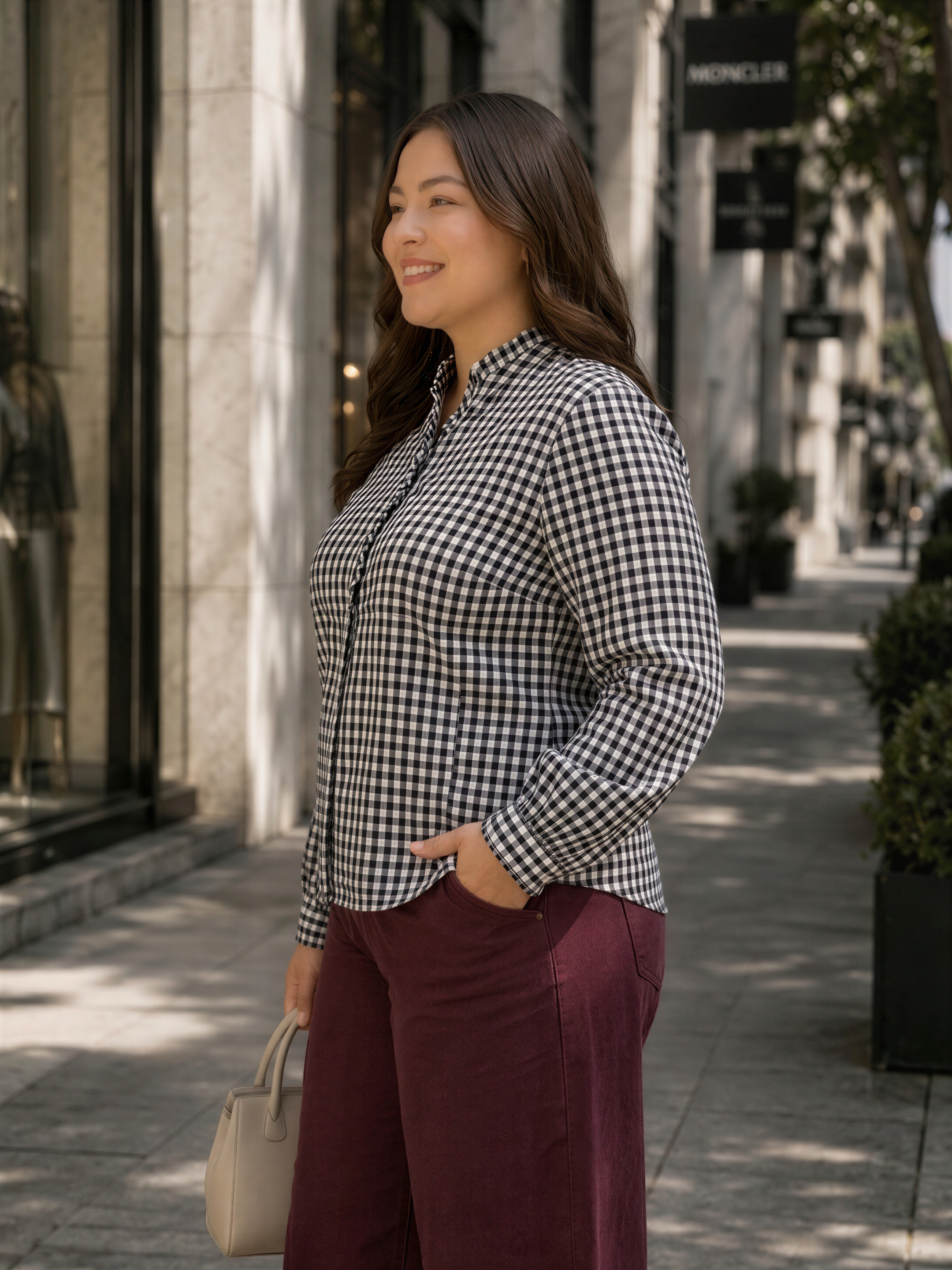 A woman wearing a black and white checkered shirt and maroon pants is standing on a sidewalk, smiling and holding a handbag.