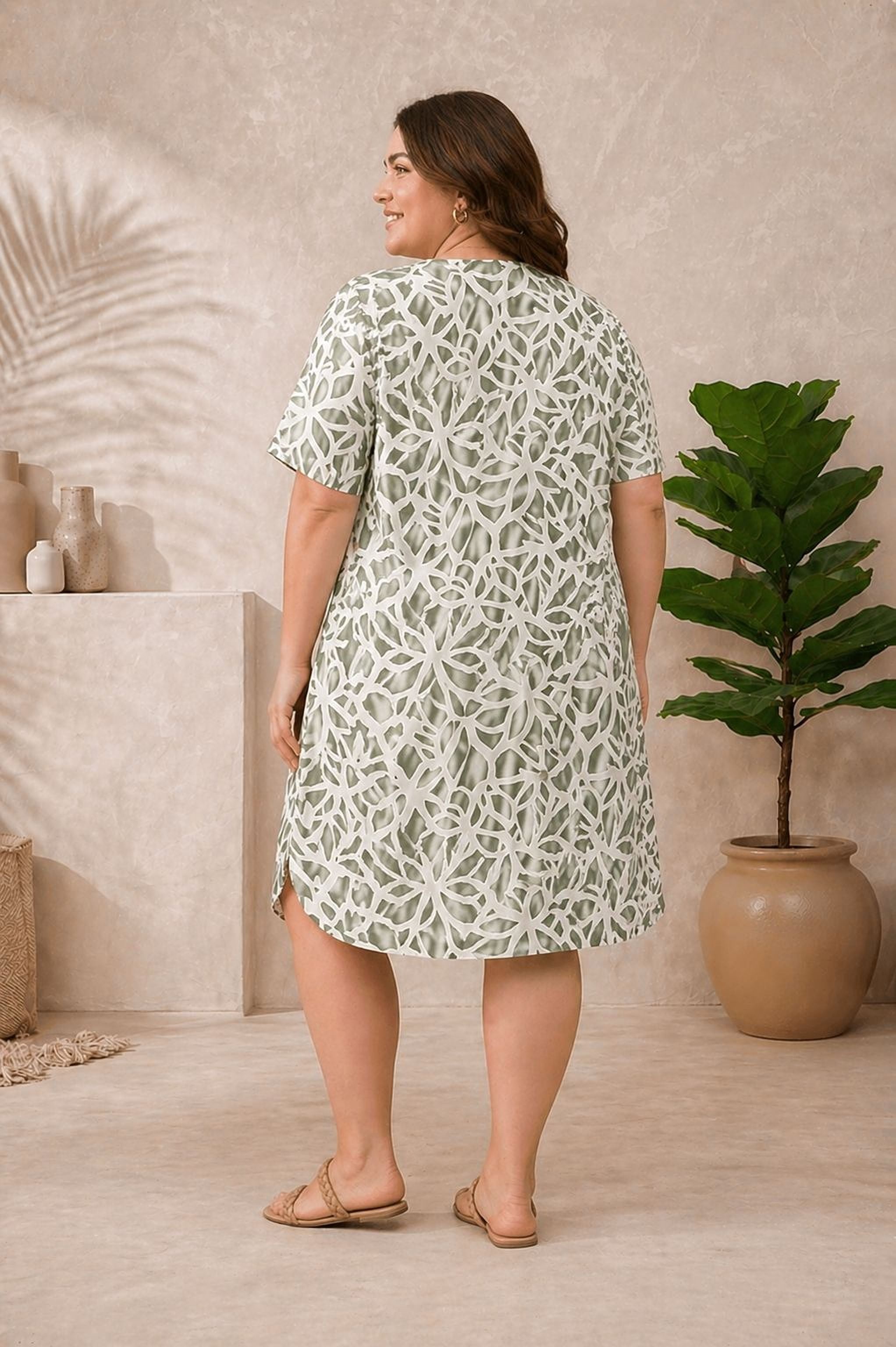 A woman wearing a white and green floral dress stands in a room with a potted plant and a shelf with vases.