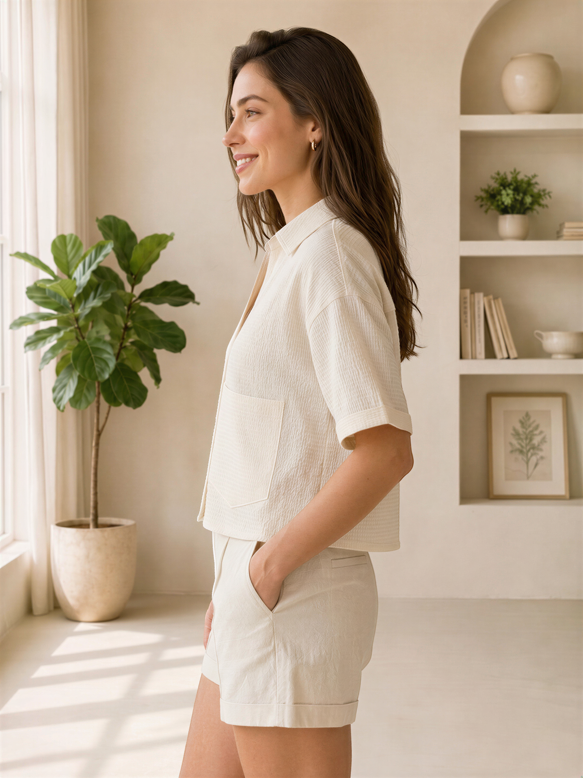 A woman in a white outfit stands in a room with a large window, smiling and looking to the side.