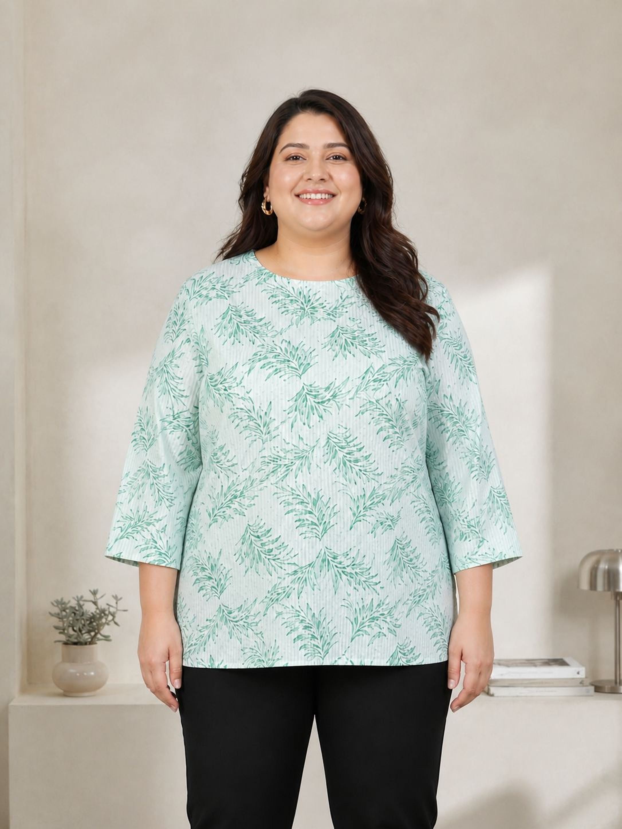 A woman wearing a green and white patterned top and black pants stands in front of a white wall, smiling at the camera.