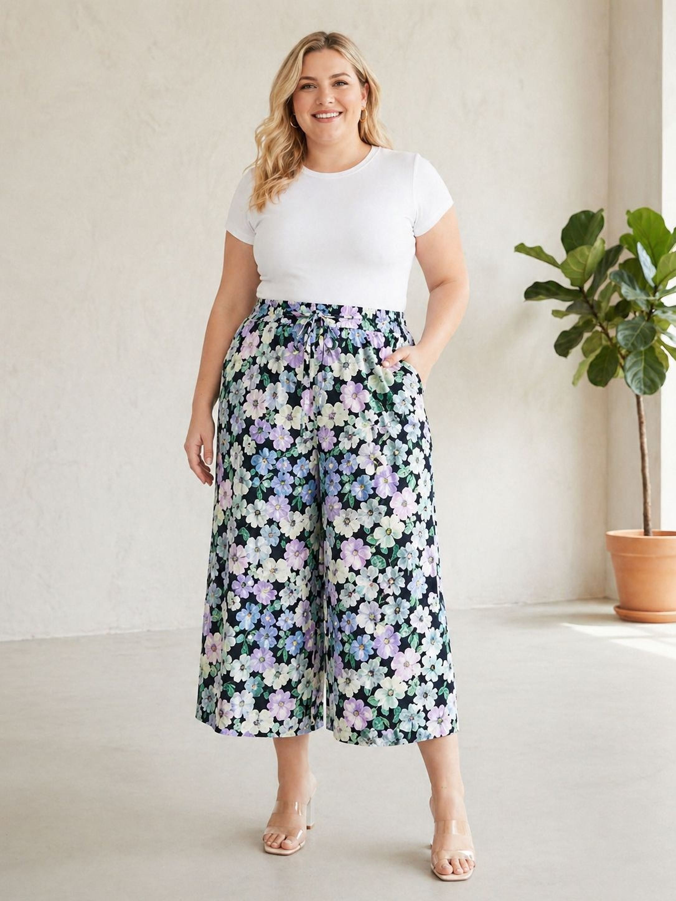 A woman wearing a white top and a floral print skirt stands in a room with a potted plant in the background.