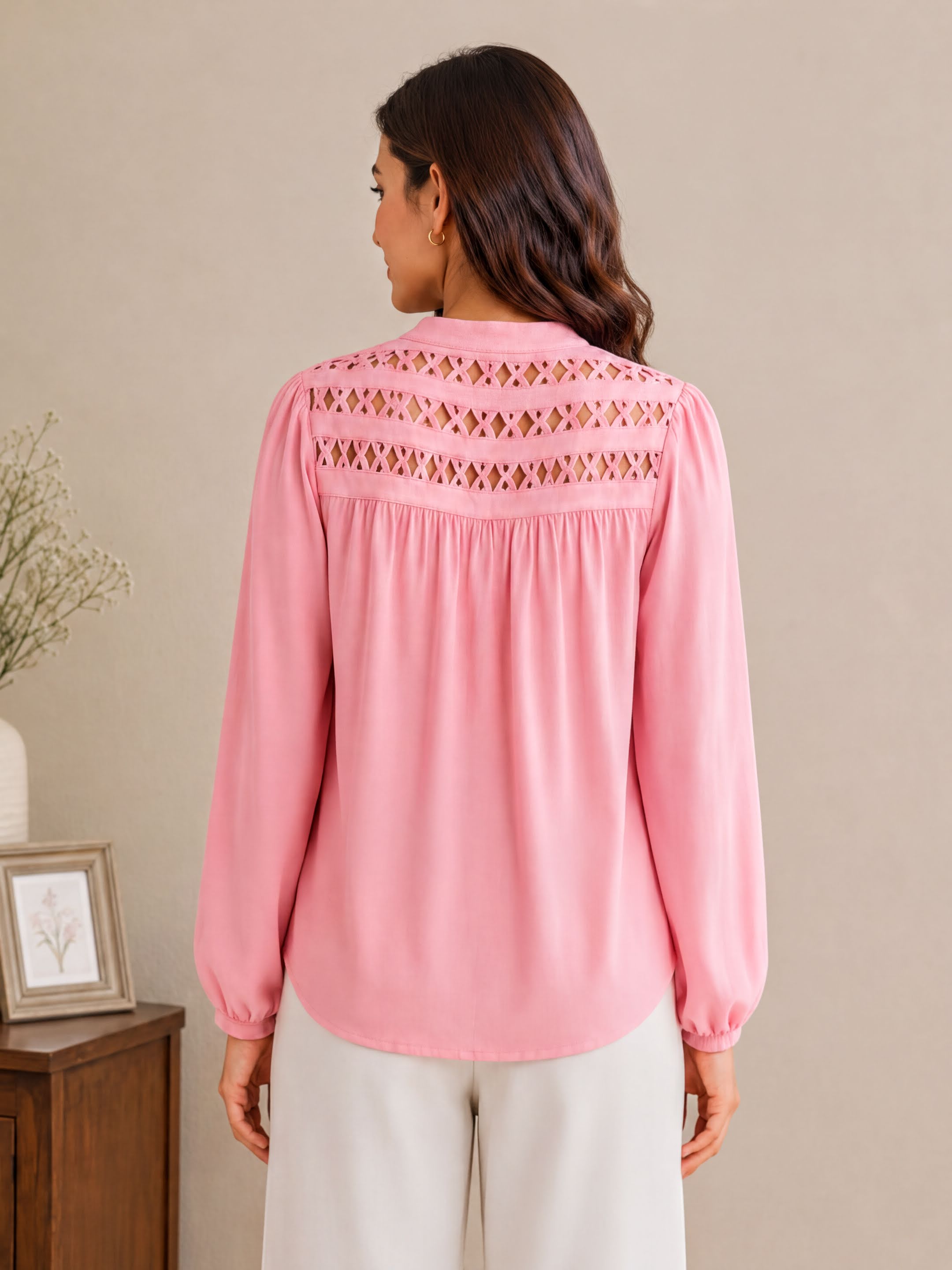 A woman wearing a pink blouse with a cutout pattern on the back, standing in front of a wooden cabinet.