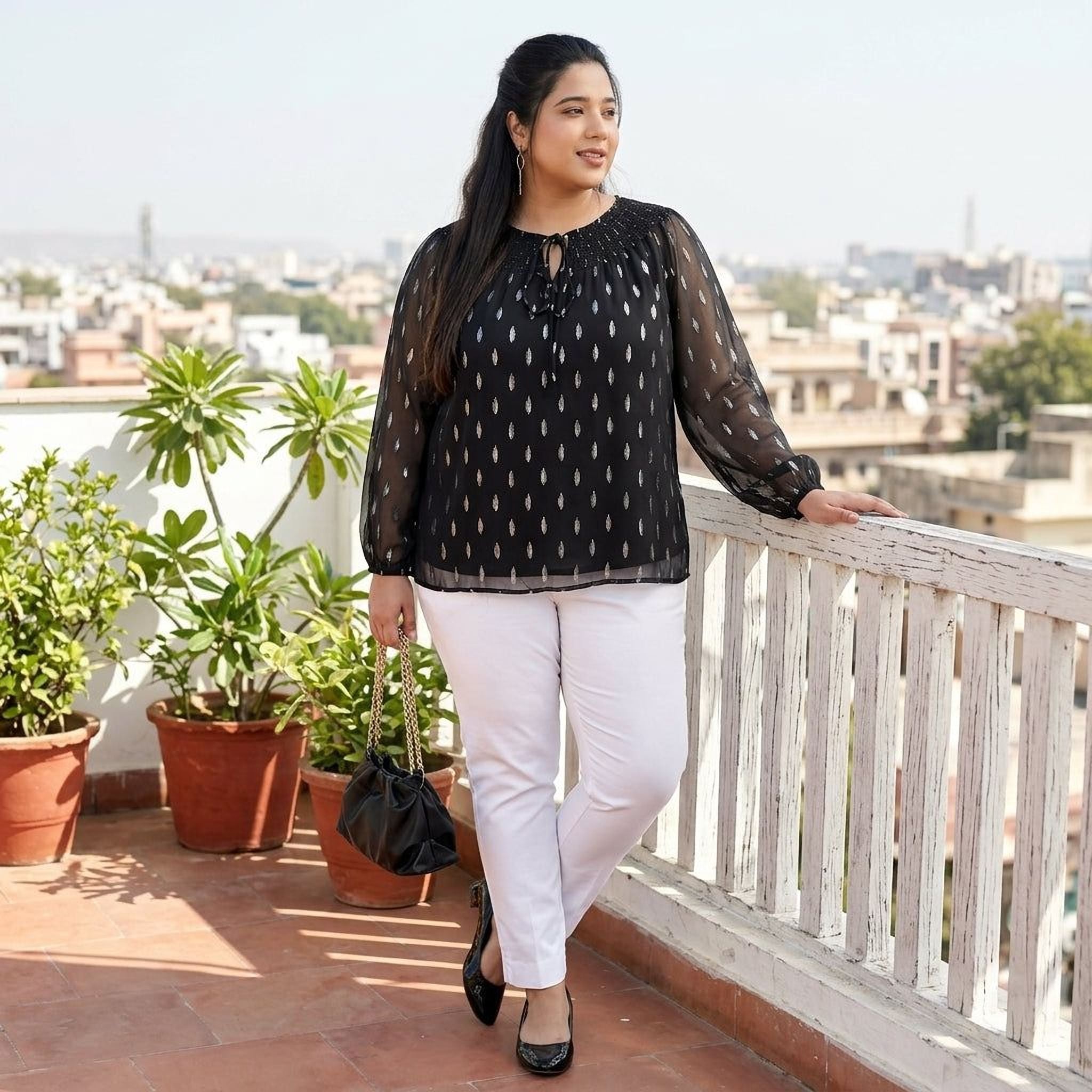 Plus size woman in black printed sheer top and white pants on rooftop, holding handbag