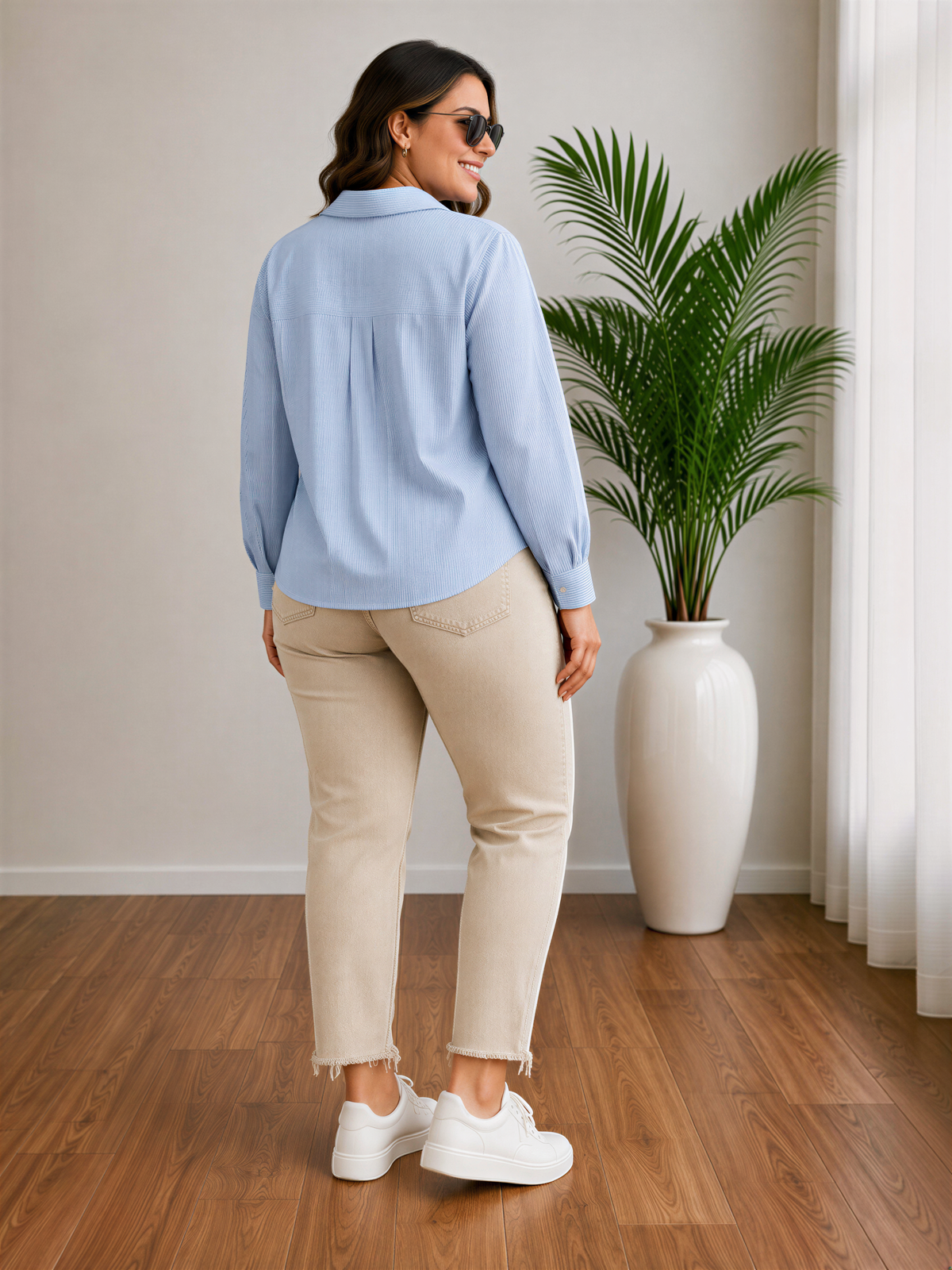 A woman wearing a light blue blouse and beige pants stands in a room with a large potted plant and a wooden floor.
