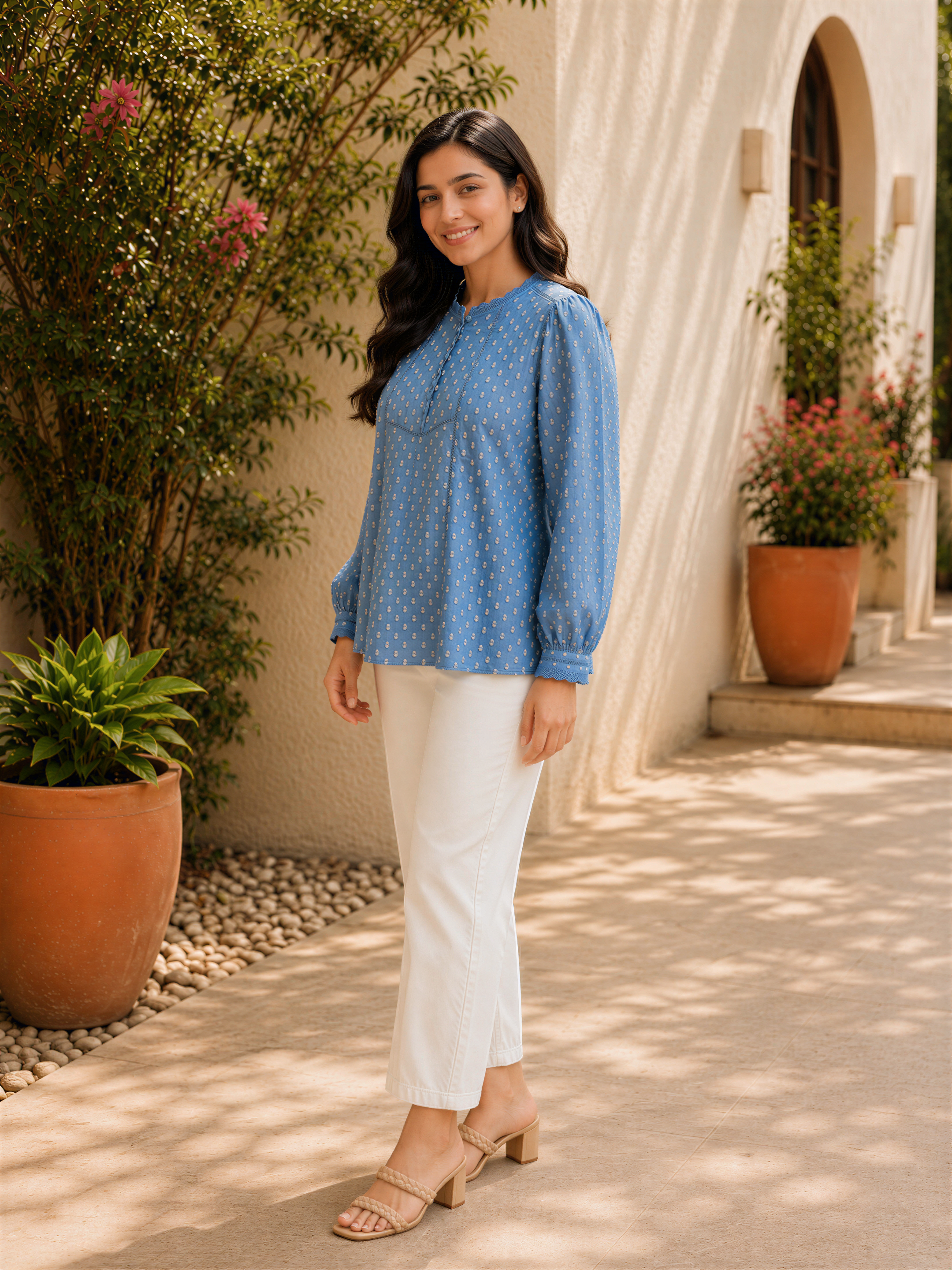 A woman in a blue polka dot blouse and white pants stands in front of a building with potted plants, smiling at the camera.