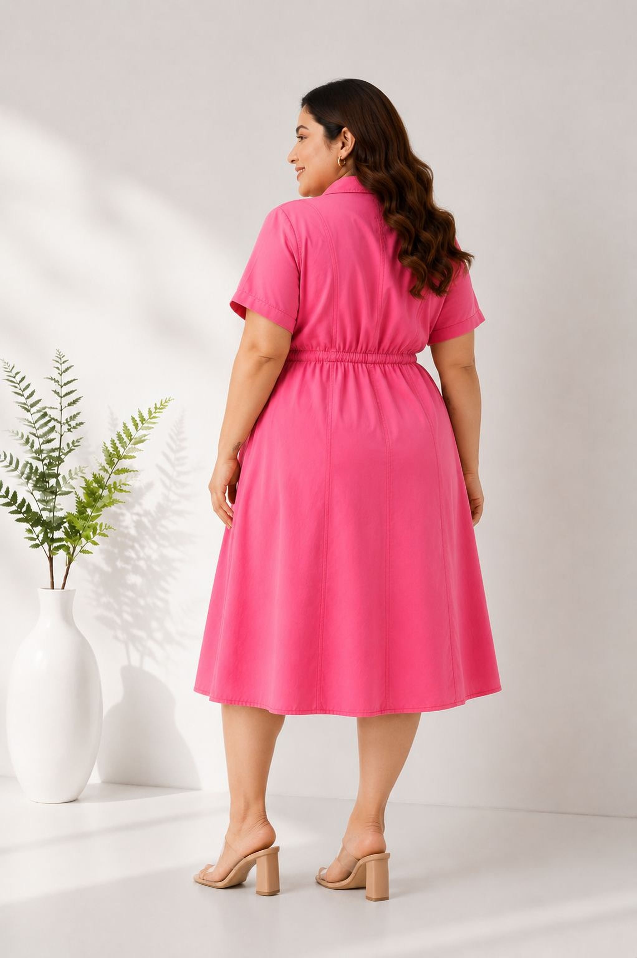 A woman wearing a bright pink dress stands in front of a white wall, with a potted plant to her left.