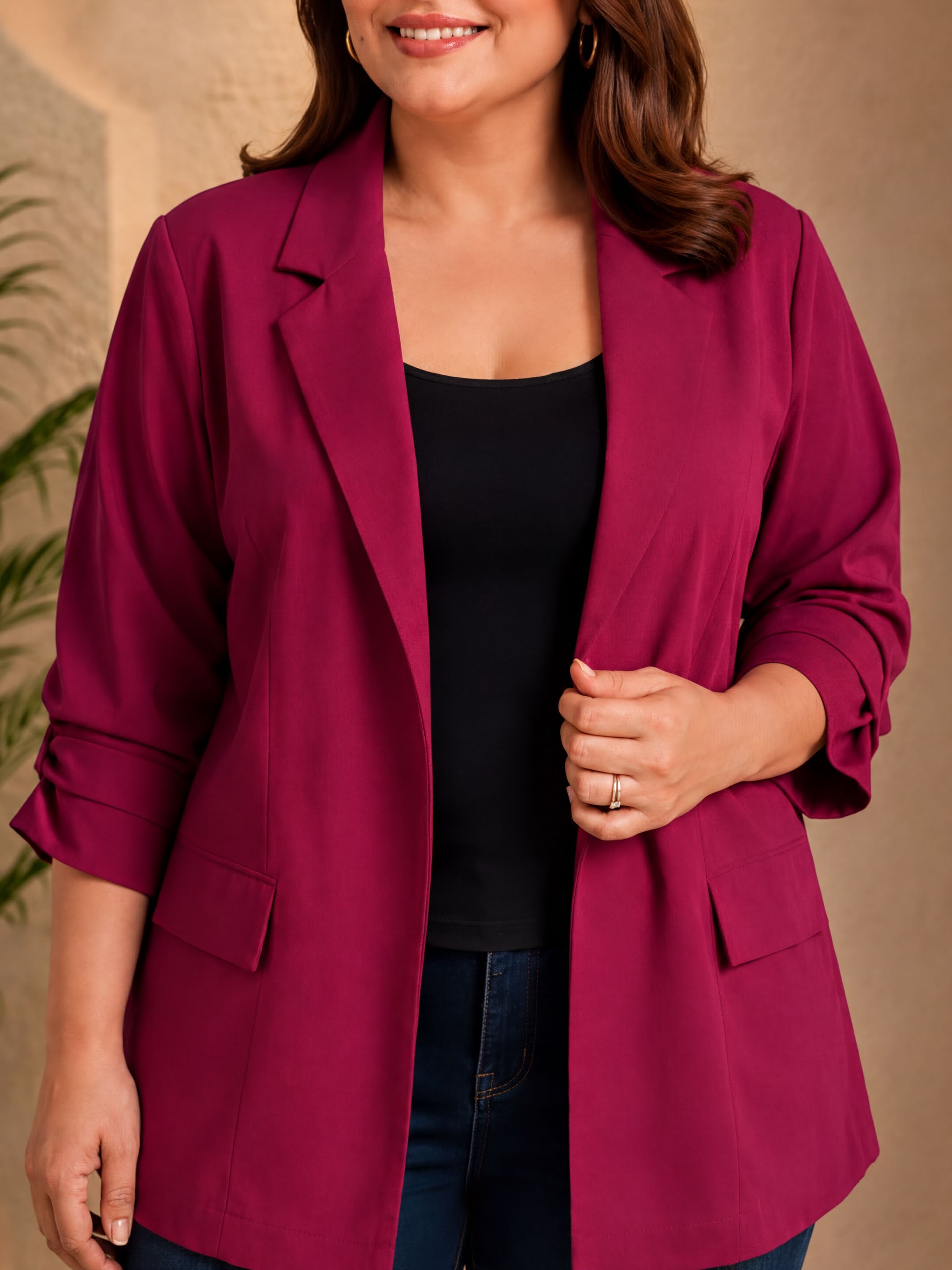 A woman wearing a burgundy blazer and black top stands in front of a beige wall, smiling at the camera.