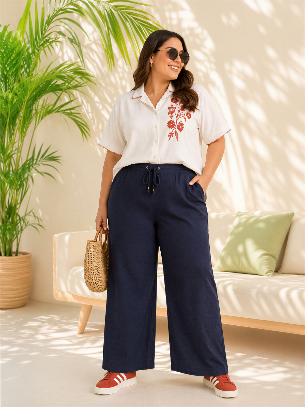 A woman wearing a white blouse with red floral embroidery, navy blue wide-leg pants, and red sneakers stands in a room with a potted plant and a couch.