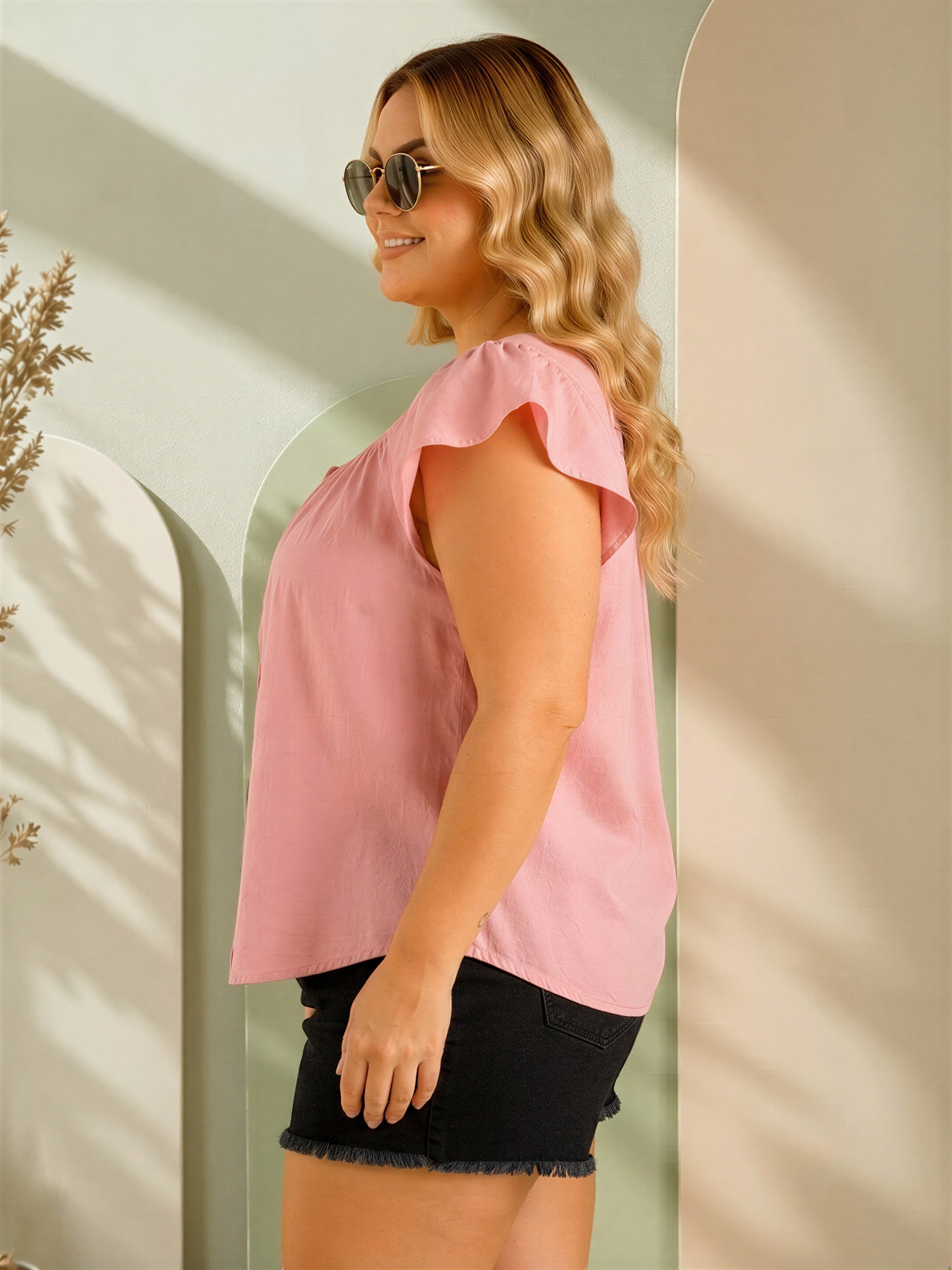 A woman with long, wavy blonde hair is standing in front of a light green wall, wearing a pink blouse and black shorts.