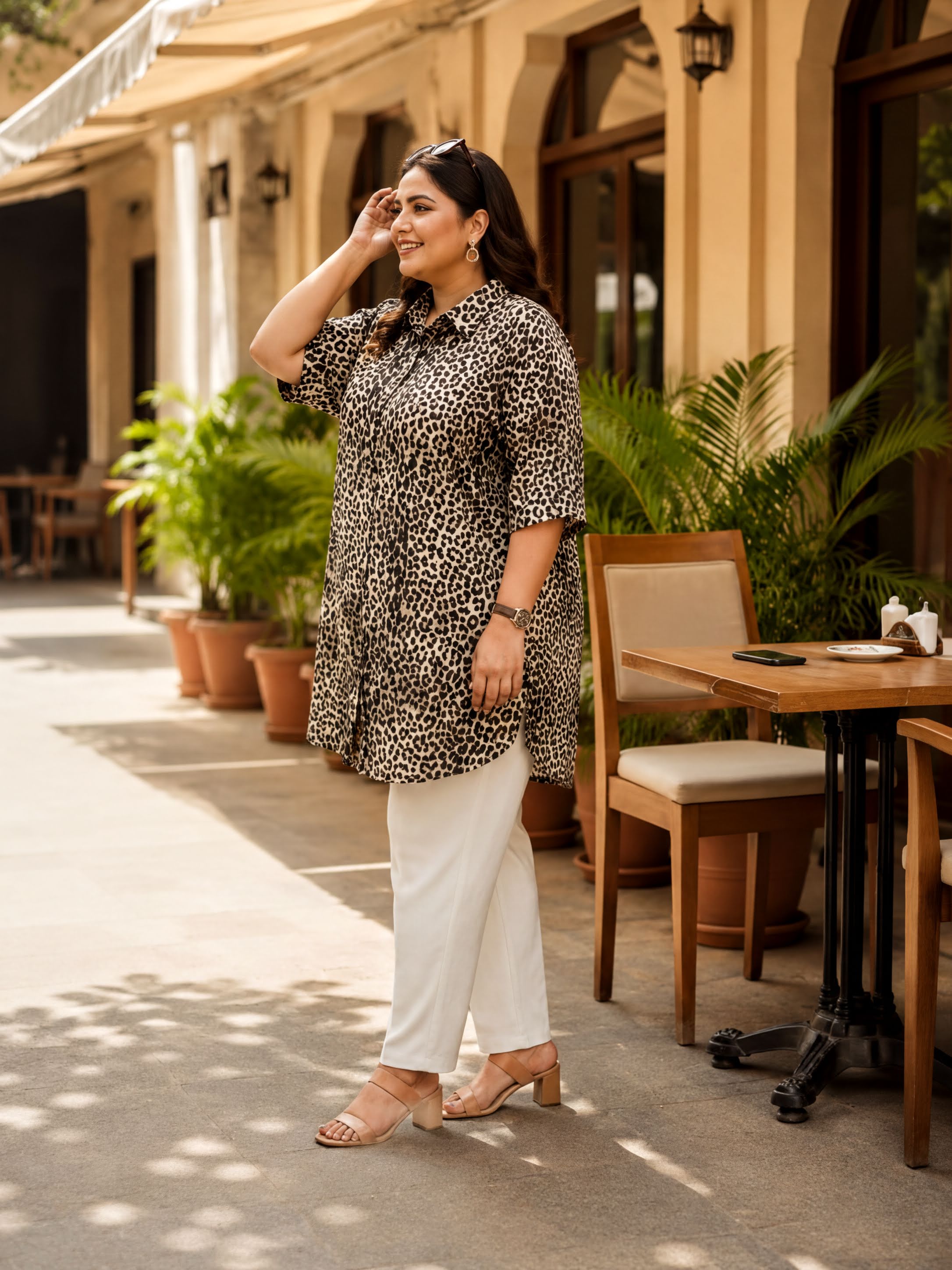 A woman wearing a leopard print dress and white pants stands on a sidewalk, smiling and looking to her left.