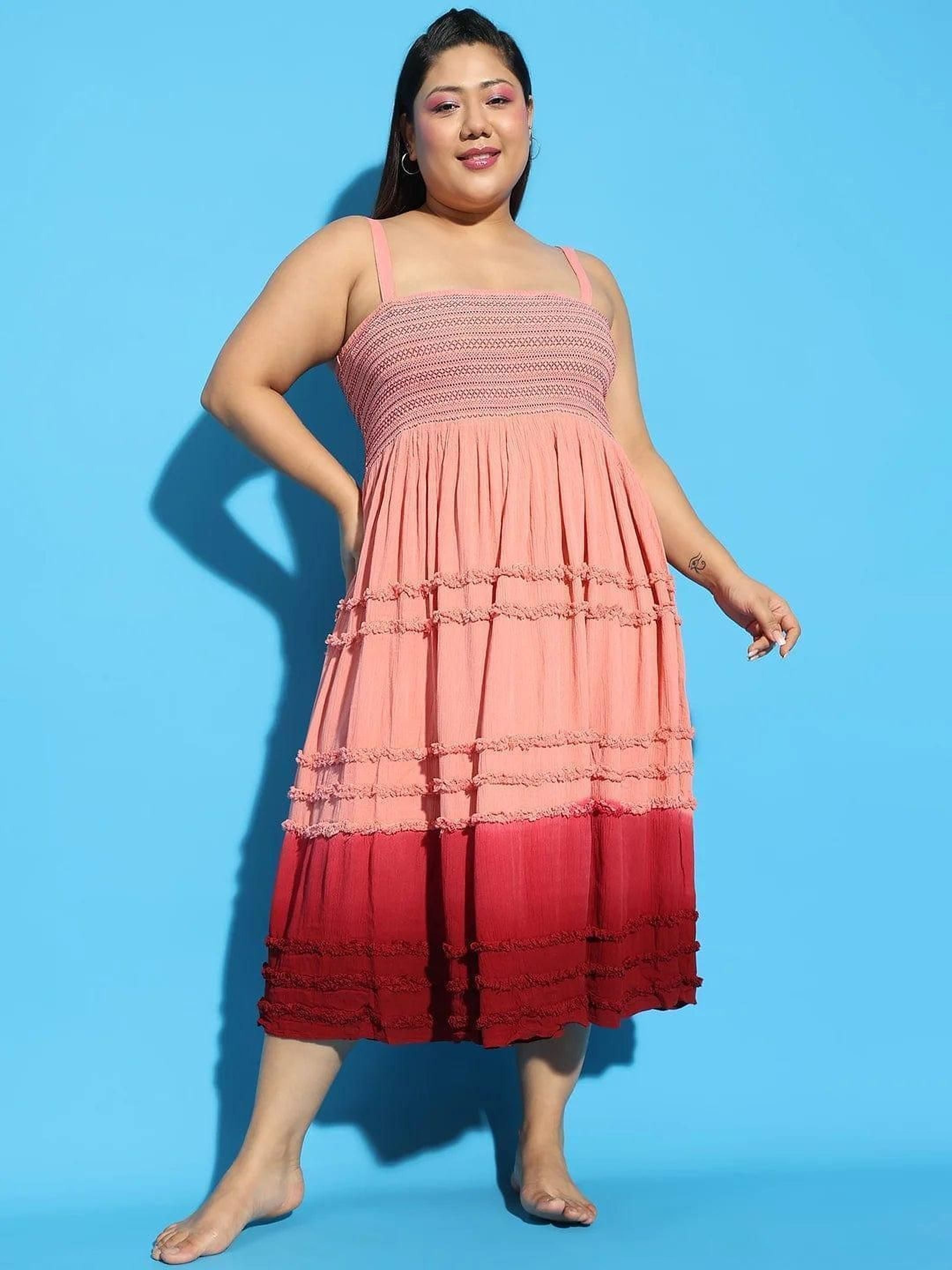 A woman wearing a pink and red tiered dress and a wide-brimmed hat is smiling and gesturing with her hand.